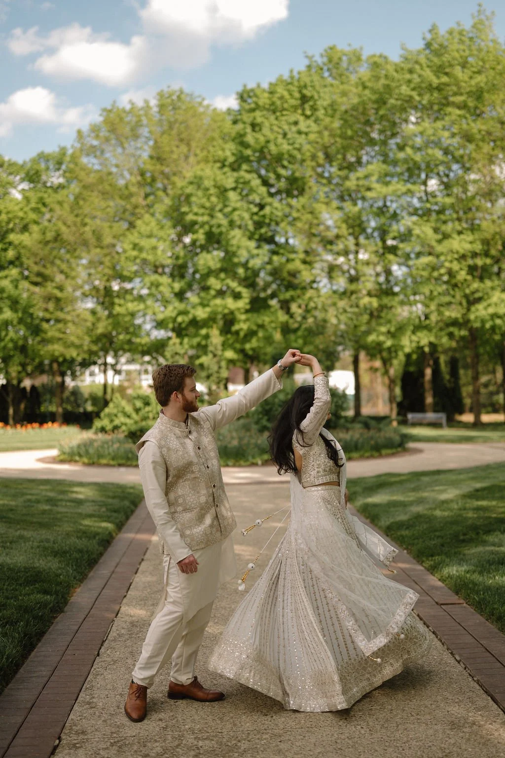 Couple in ivory outfits dancing on a garden path as she twirls, with her skirt and dupatta flowing under green trees.
