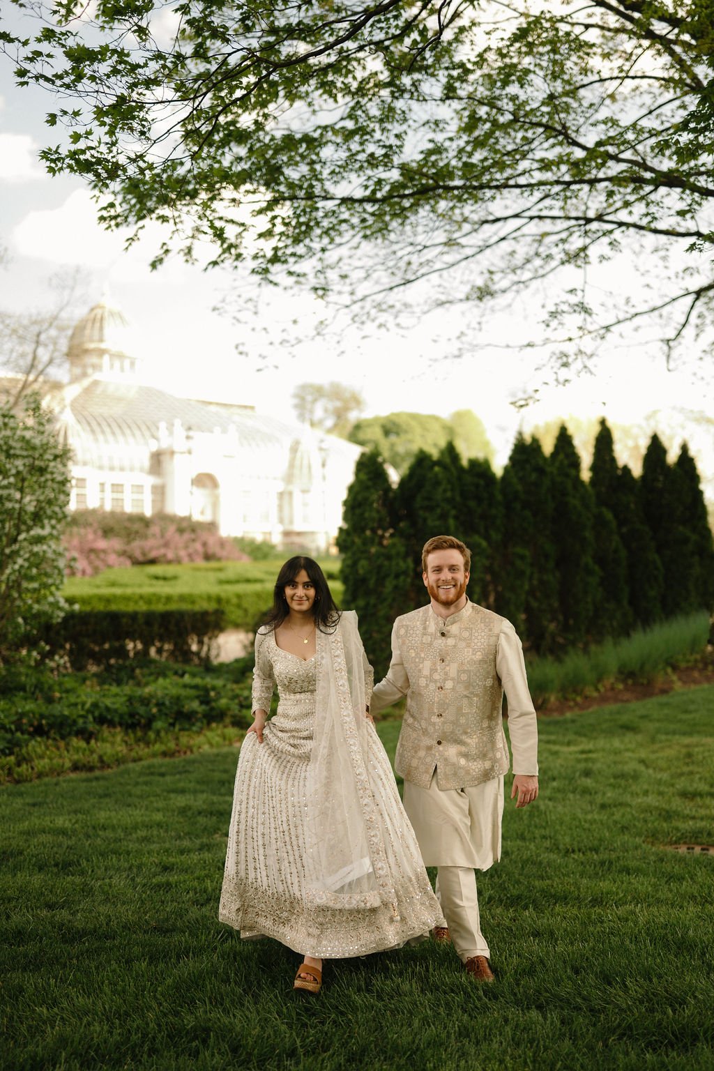 Engagement photo ideas with a laughing couple in ivory outfits walking toward the camera on a lawn, with a glass conservatory behind them.