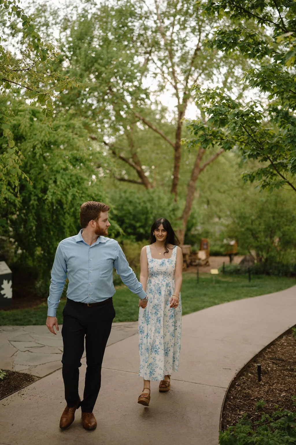 Man glancing back as he walks hand in hand with his smiling fiancée in a blue floral dress along a curved garden path.