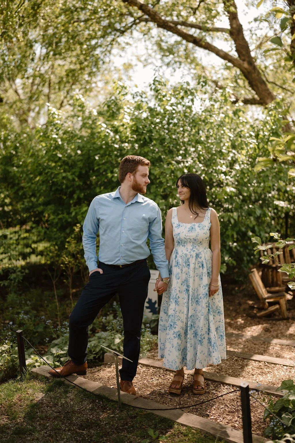 Engagement photo ideas with a couple holding hands on a wood chip path in a sunny garden, smiling at each other.