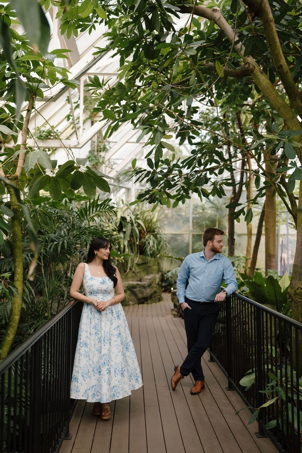 Couple in a blue floral dress and light blue button up leaning on black railings on a wooden walkway, glancing in opposite directions under tropical trees.