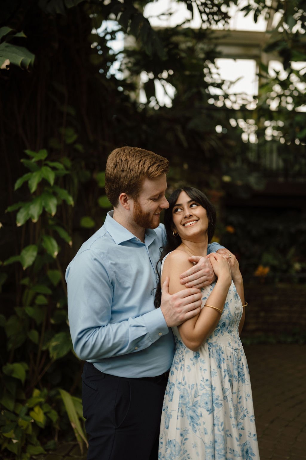Engagement photo ideas with a woman in a blue floral dress smiling up at her fiancé as he wraps his arms around her in a dark, leafy greenhouse.