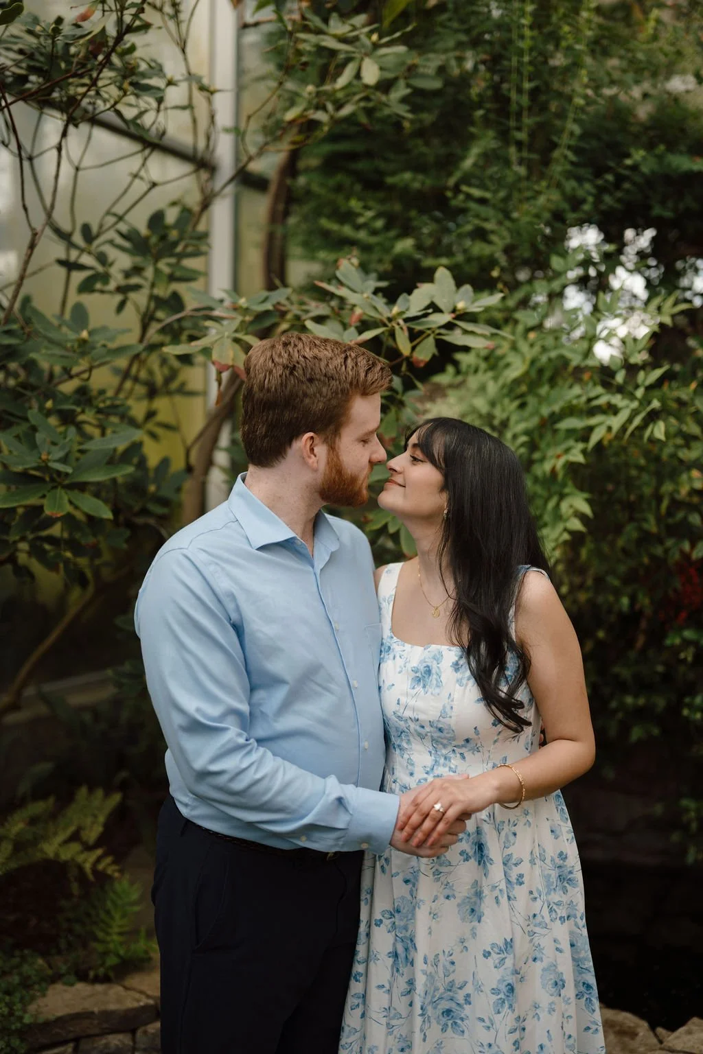 Couple in blue standing nose to nose and holding hands in front of rhododendron bushes, with her engagement ring in view.