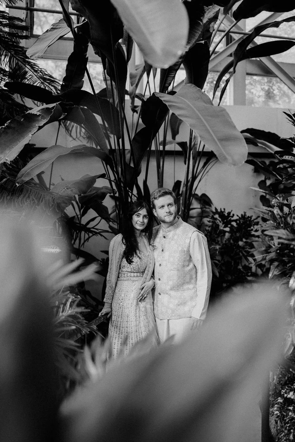 Black and white photo of a couple standing close together, framed by large tropical leaves in a conservatory.