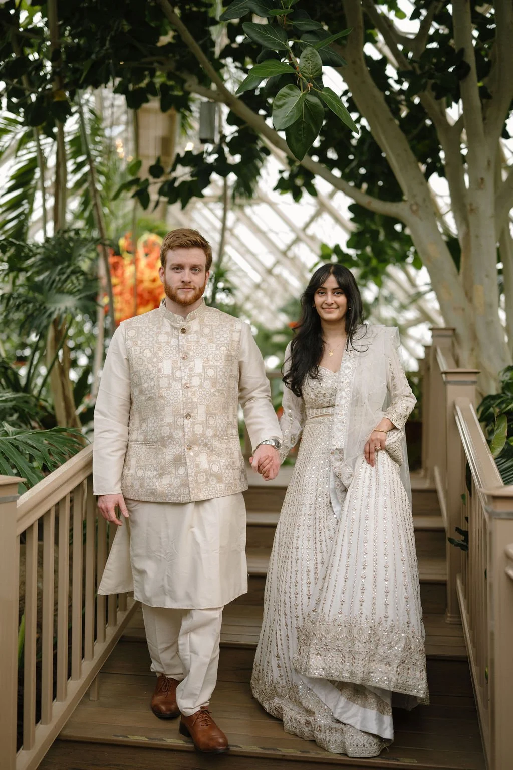 Engagement photo ideas featuring a smiling couple in coordinating ivory outfits walking hand in hand down wooden steps inside a greenhouse.
