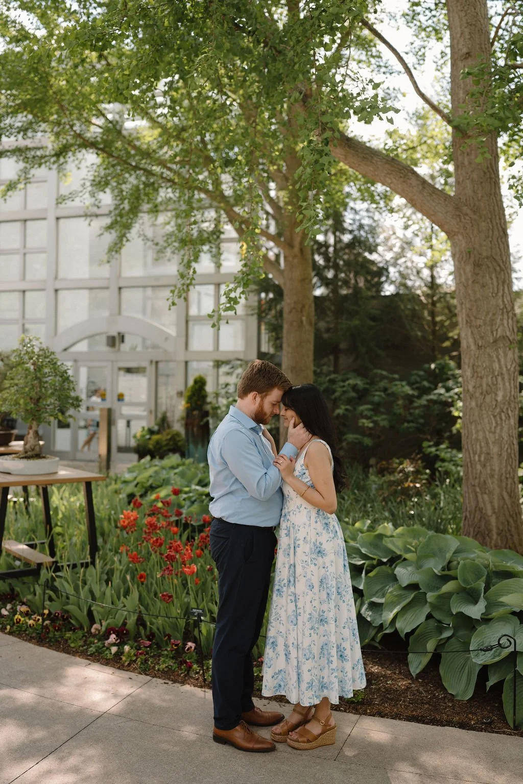 Couple touching foreheads on a garden path beside red tulips and large hosta leaves, with a glass conservatory behind them.