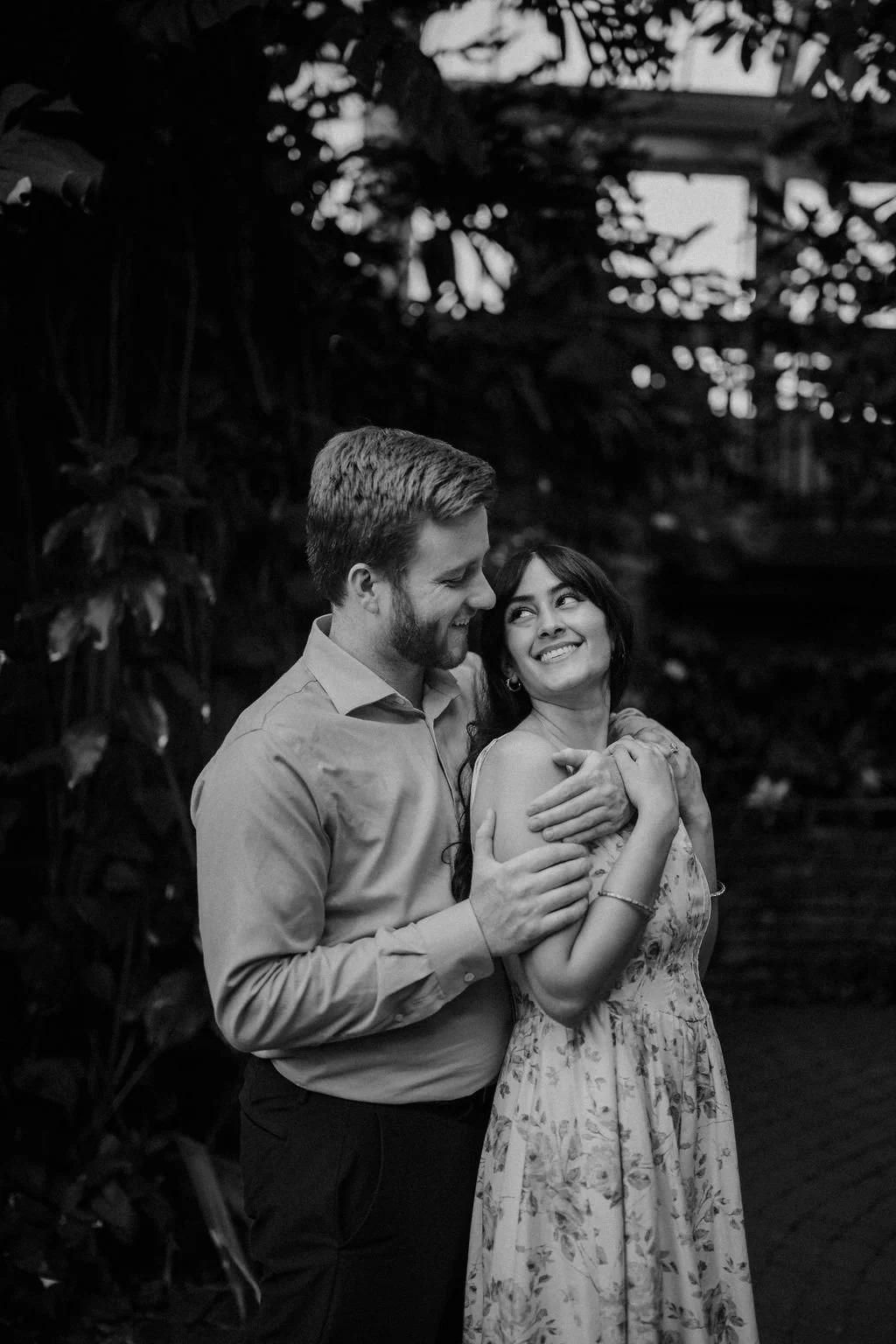 Engagement photo ideas for a sweet candid moment, with a woman in a floral dress looking up and smiling as her fiancé hugs her from behind in a black and white photo.
