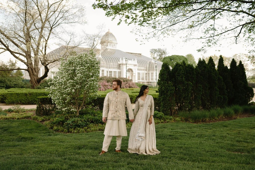 Engagement photo ideas with a couple holding hands on a green lawn, with a glass conservatory and white flowering tree behind them.