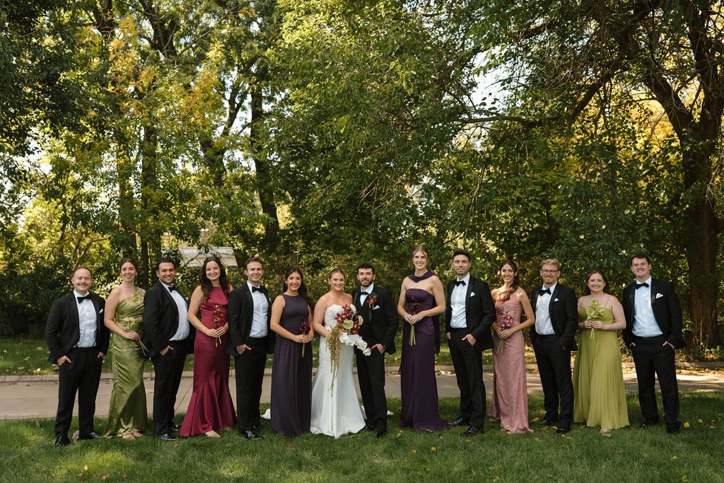 Bride laughing with six bridesmaids in burgundy, olive, navy, and dusty rose gowns under a canopy of green trees