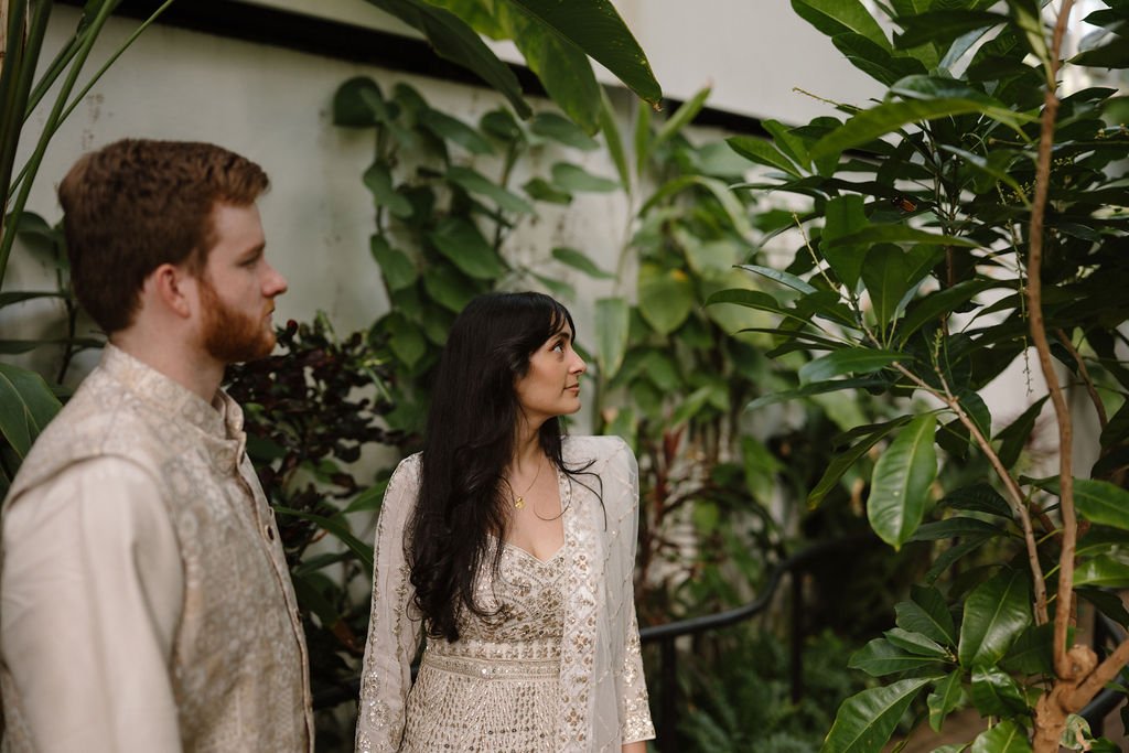 Engagement photo ideas that feel quiet and candid, with a couple in ivory outfits looking off to the side among green leaves in a conservatory.