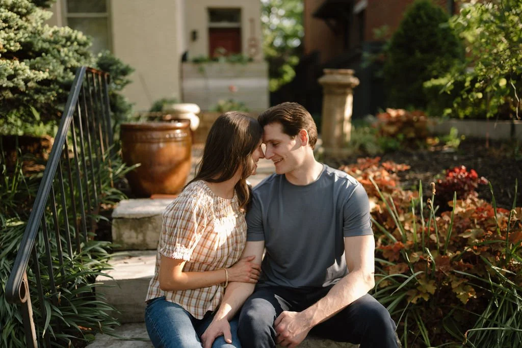 Couple sitting on stone garden steps, foreheads touching, surrounded by colorful plants and potted pottery.
