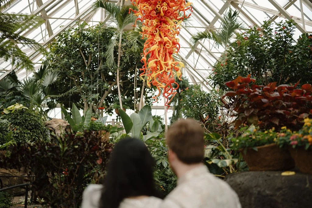 Couple seen from behind looking up at a large orange and yellow glass sculpture hanging inside a greenhouse.