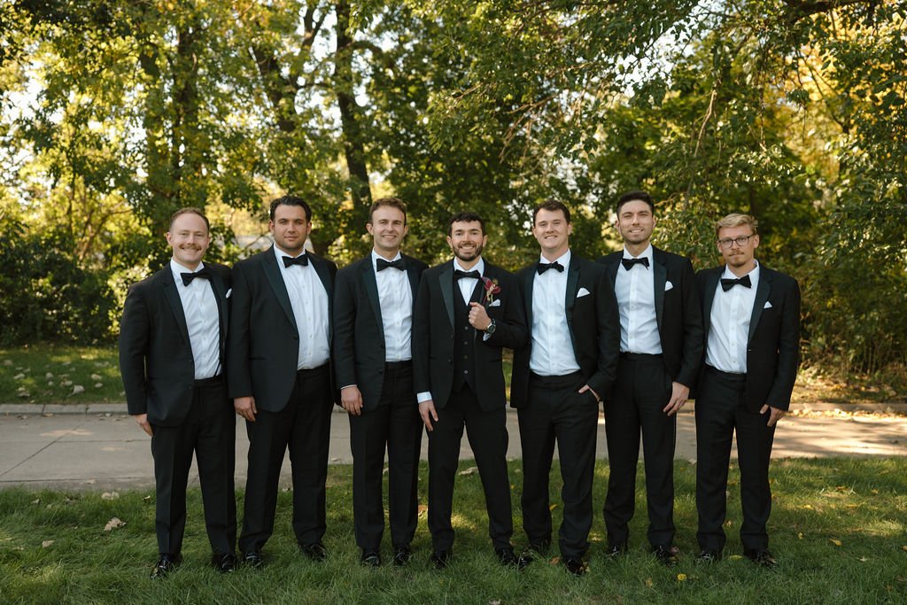 Seven groomsmen in black tuxedos and bow ties standing in a row under trees with the groom in the center