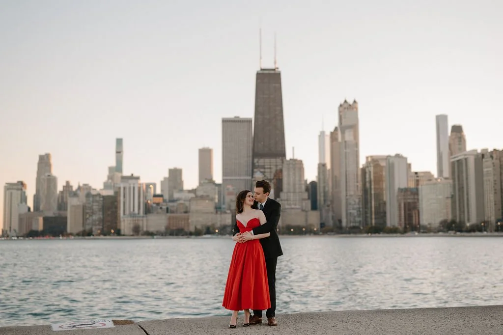 Chicago engagement photos at sunset, with a couple embracing along Lake Michigan, the skyline behind them and her in a red dress