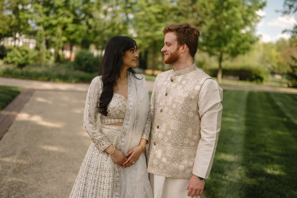 Engagement photo ideas with a couple in ivory traditional outfits smiling at each other on a sunny garden path lined with trees and grass.