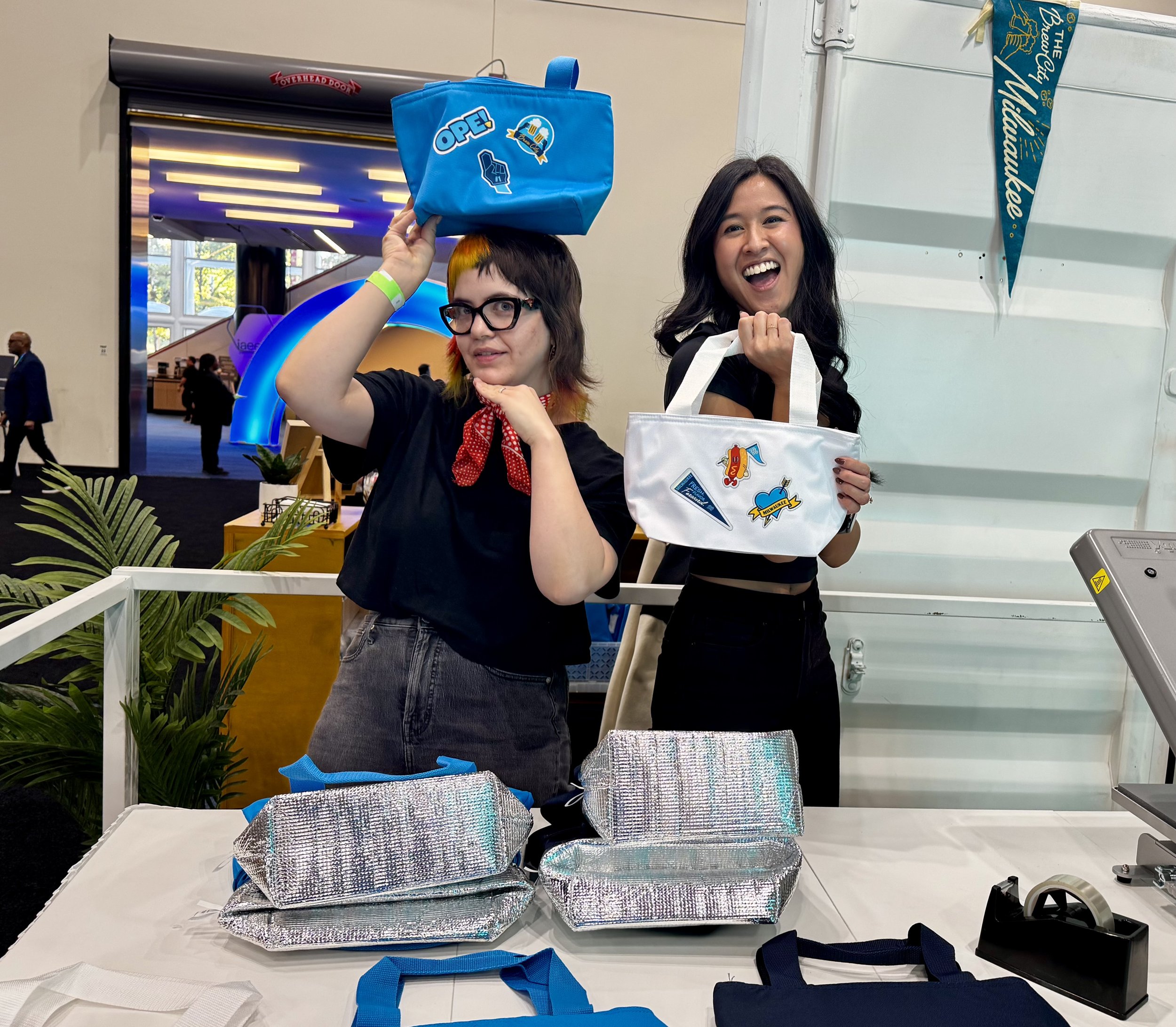 Gabby and Maddie goof while modeling the cooler tote bags they will be heat pressing live forVisit Milwaukee's booth at Expo Expo 2025