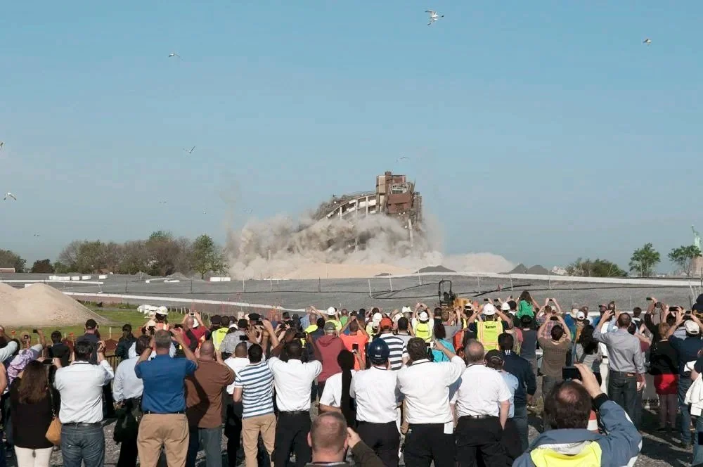 A building collapsing into dust and debris while a crowd of onlookers and reporters watch and take photos nearby.