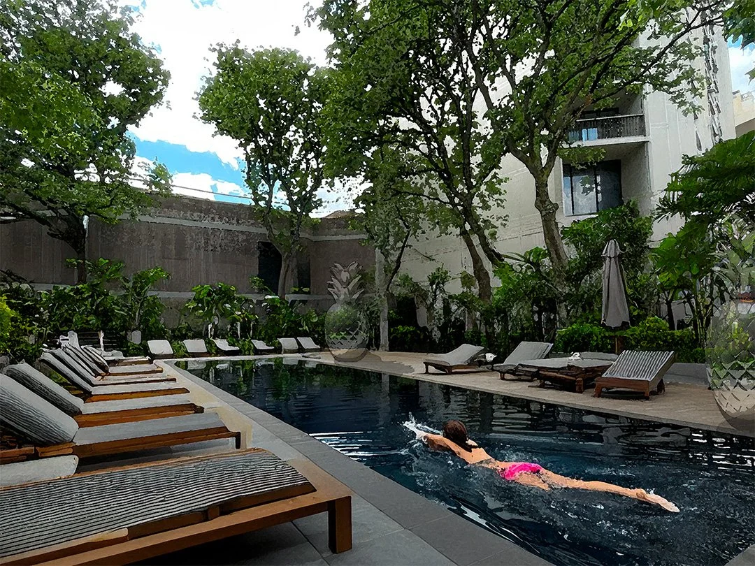 A woman swimming in a private outdoor pool surrounded by lounge chairs and lush green trees.