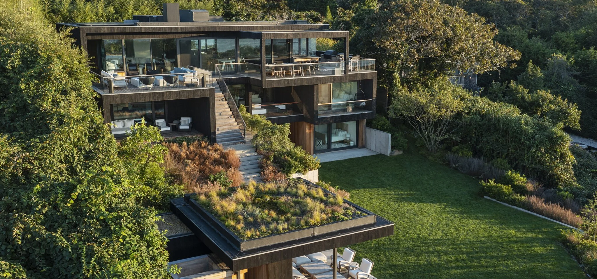 Modern multi-level house surrounded by lush greenery, featuring large glass windows, balconies, and a rooftop garden, with an open green lawn and trees in the foreground.