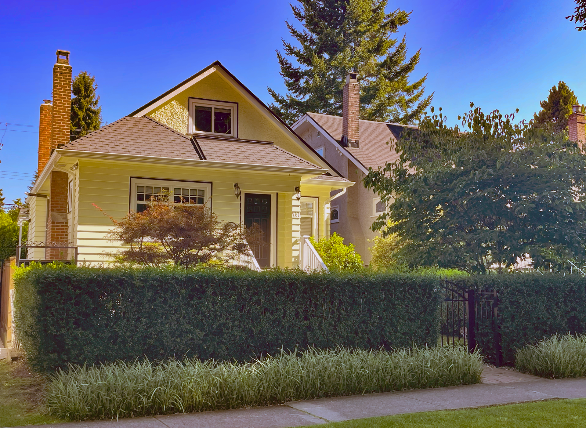 A light yellow house with white trim and a dark door, surrounded by green bushes and trees, under a blue sky.