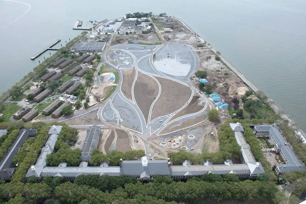 Aerial view of a large park area under construction next to a body of water, with winding pathways, some buildings, and trees surrounding the site.
