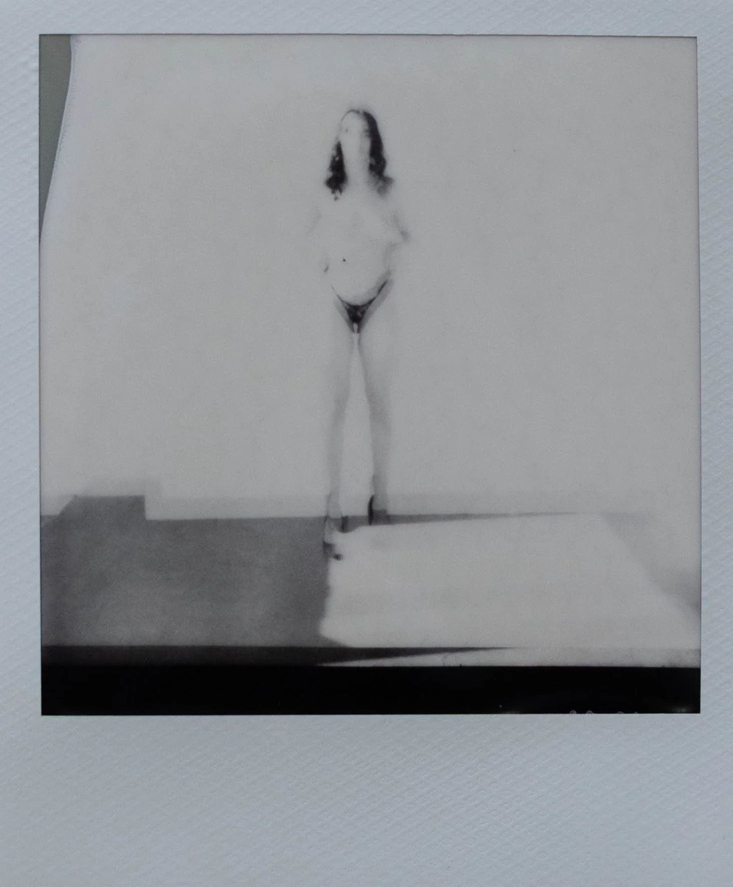 Black and white polaroid of a woman standing in black heals and black undies covering her breasts with her hands