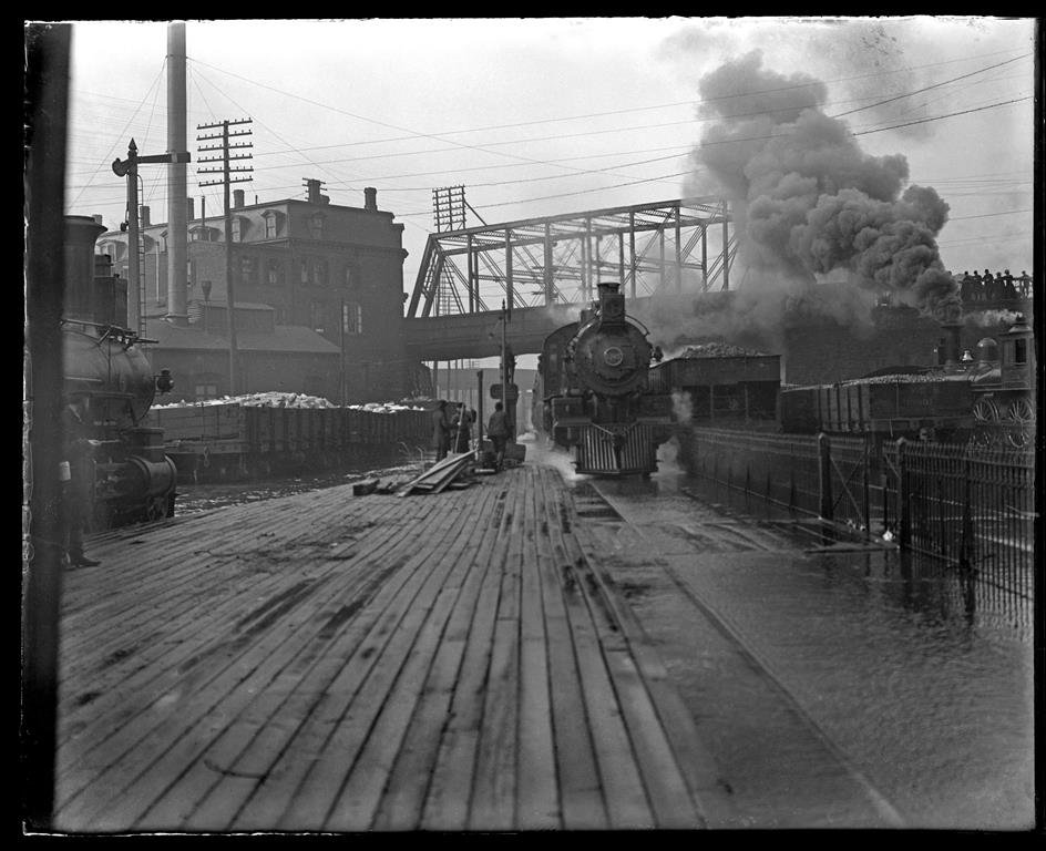 A locomotive with a bridge and a factory in the background
