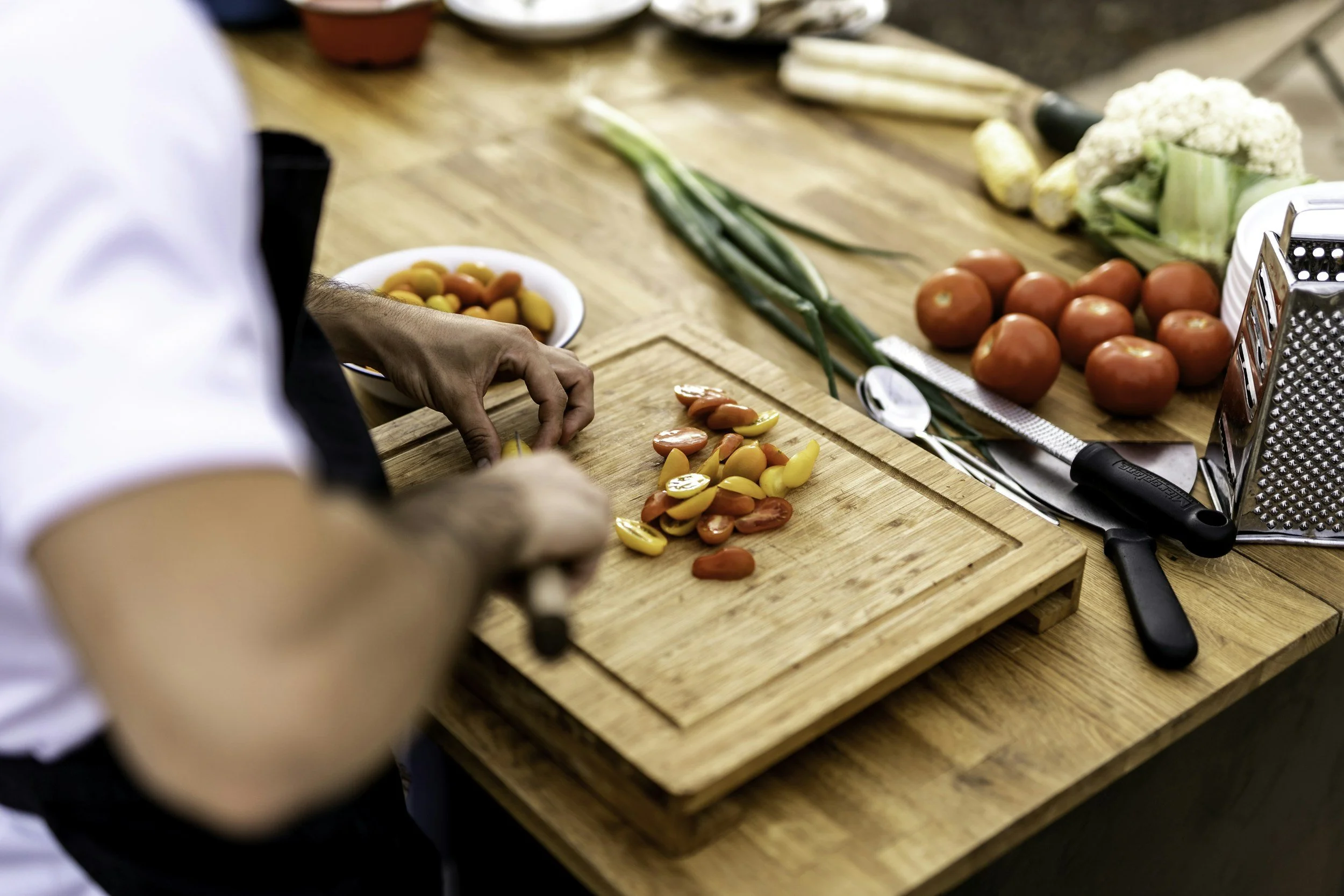 Person slicing cherry tomatoes on a wooden cutting board in a kitchen. Various vegetables including tomatoes, cauliflower, and green onions on the countertop.