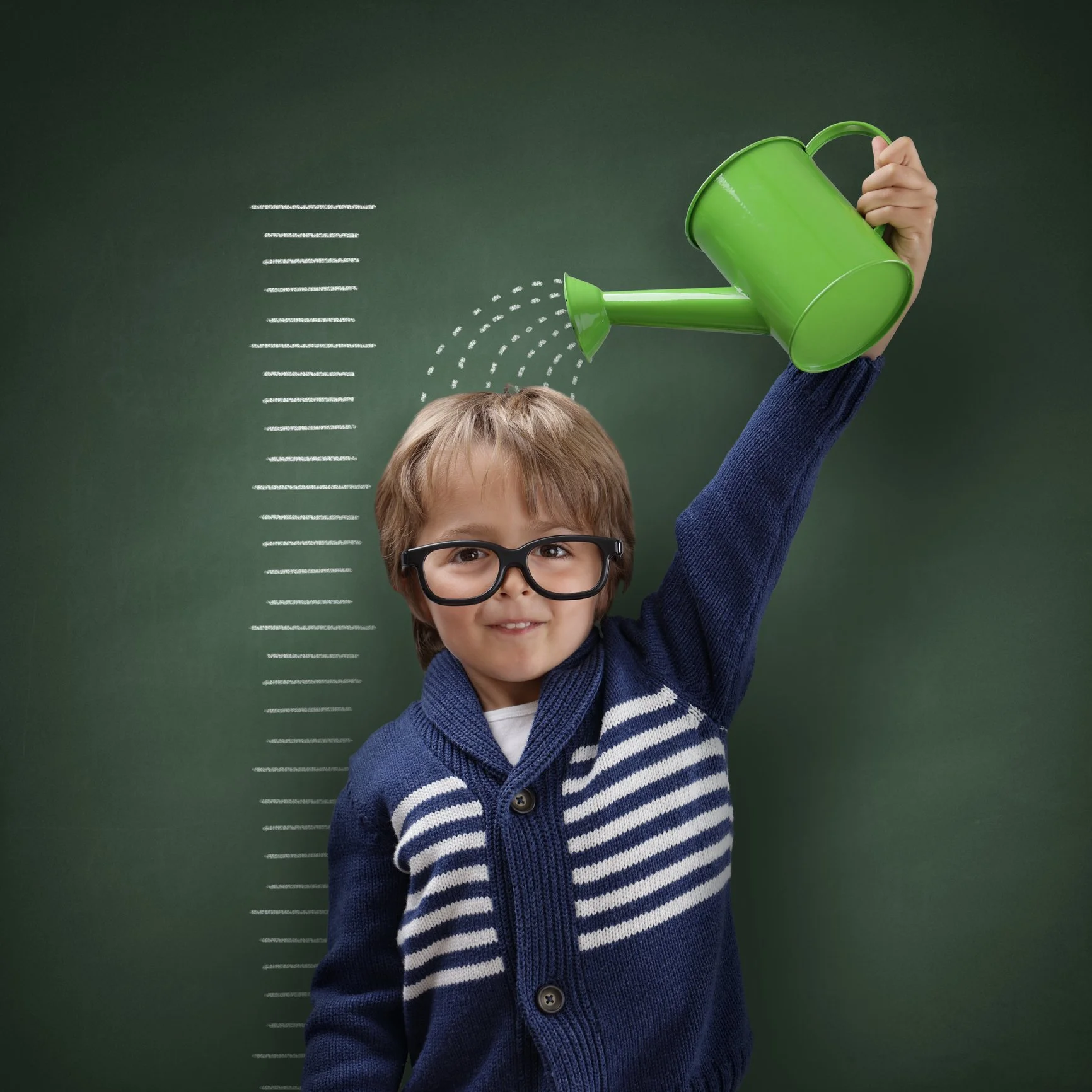 A young boy with glasses smiling and raising his arm, holding a green watering can over his head in front of a green chalkboard with height lines and chalk-drawn water spray.