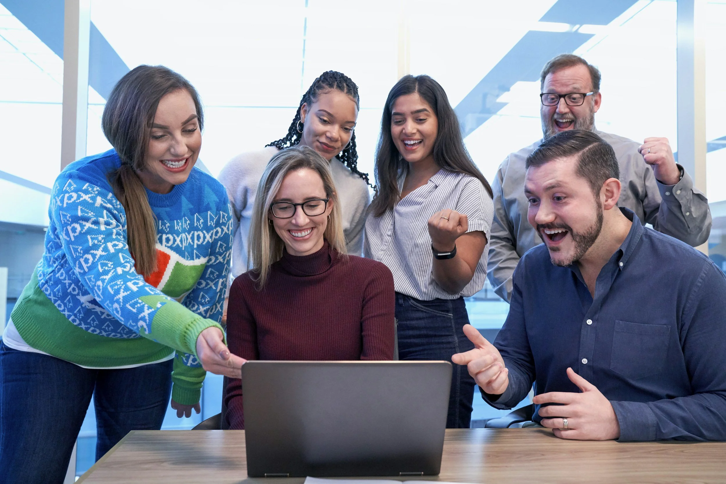 Group of six people gathered around a laptop, smiling and celebrating indoors.