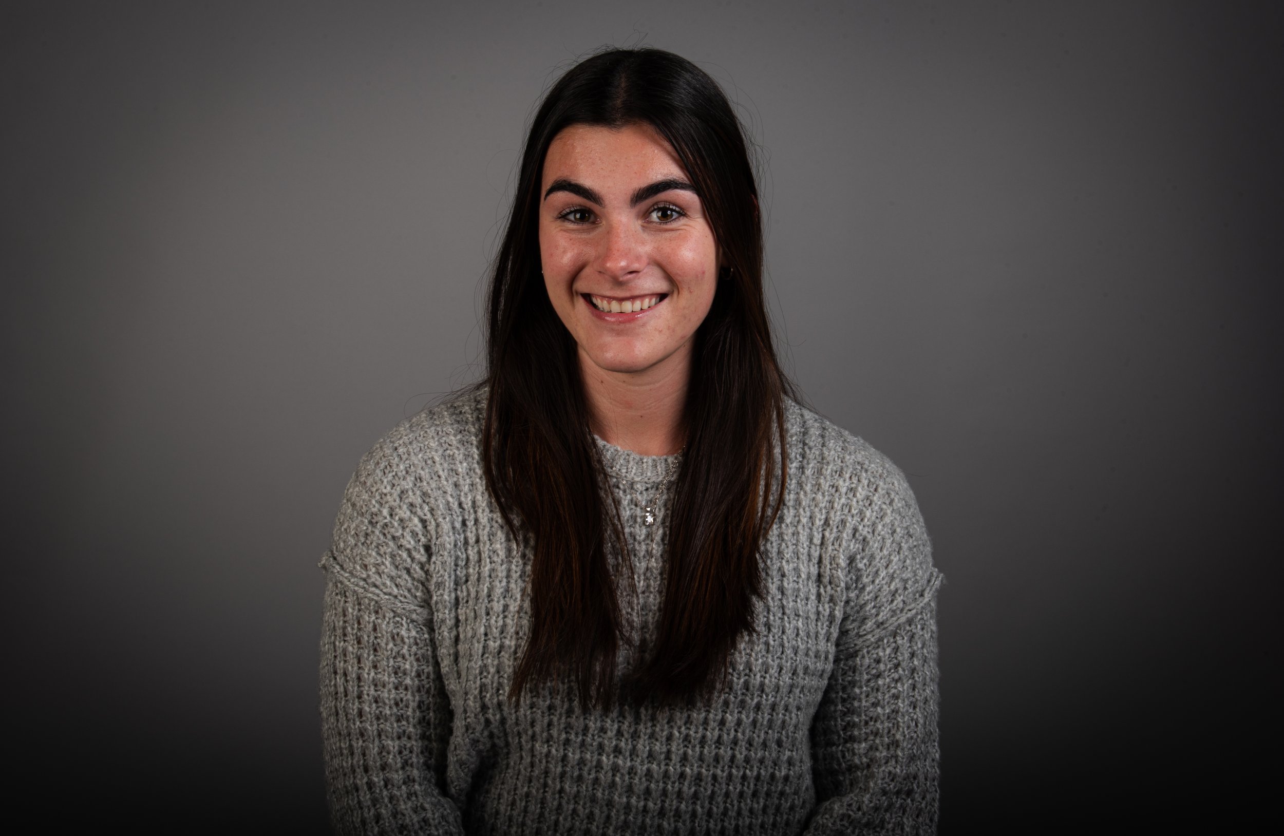A young woman with long dark hair, smiling, wearing a gray knit sweater, sitting against a plain gray background.