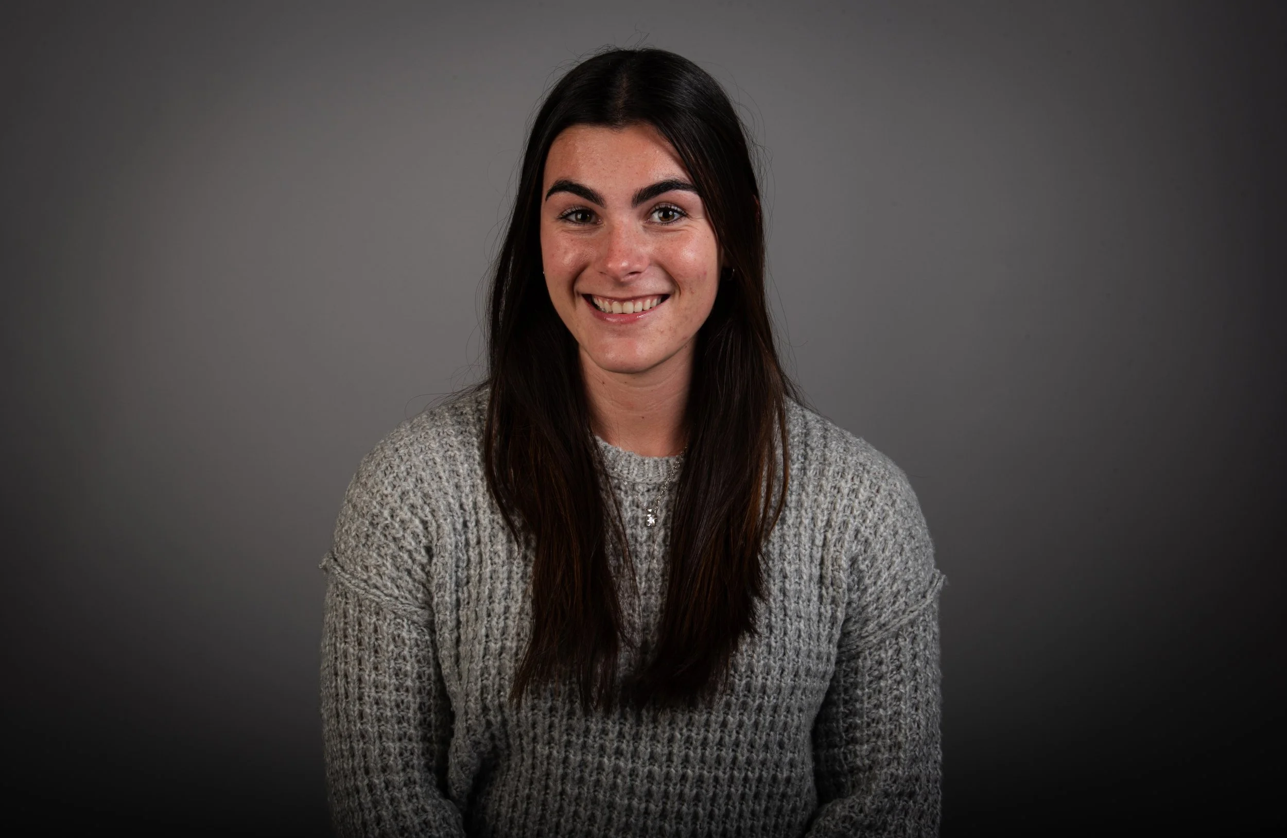 Young woman with long dark hair, smiling, wearing a gray knit sweater, seated against a plain gray background.