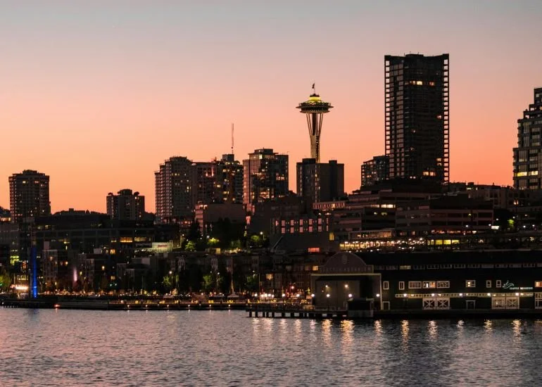 Seattle skyline at sunset viewed across the waterfront with the Space Needle, city lights, and calm water in the foreground.