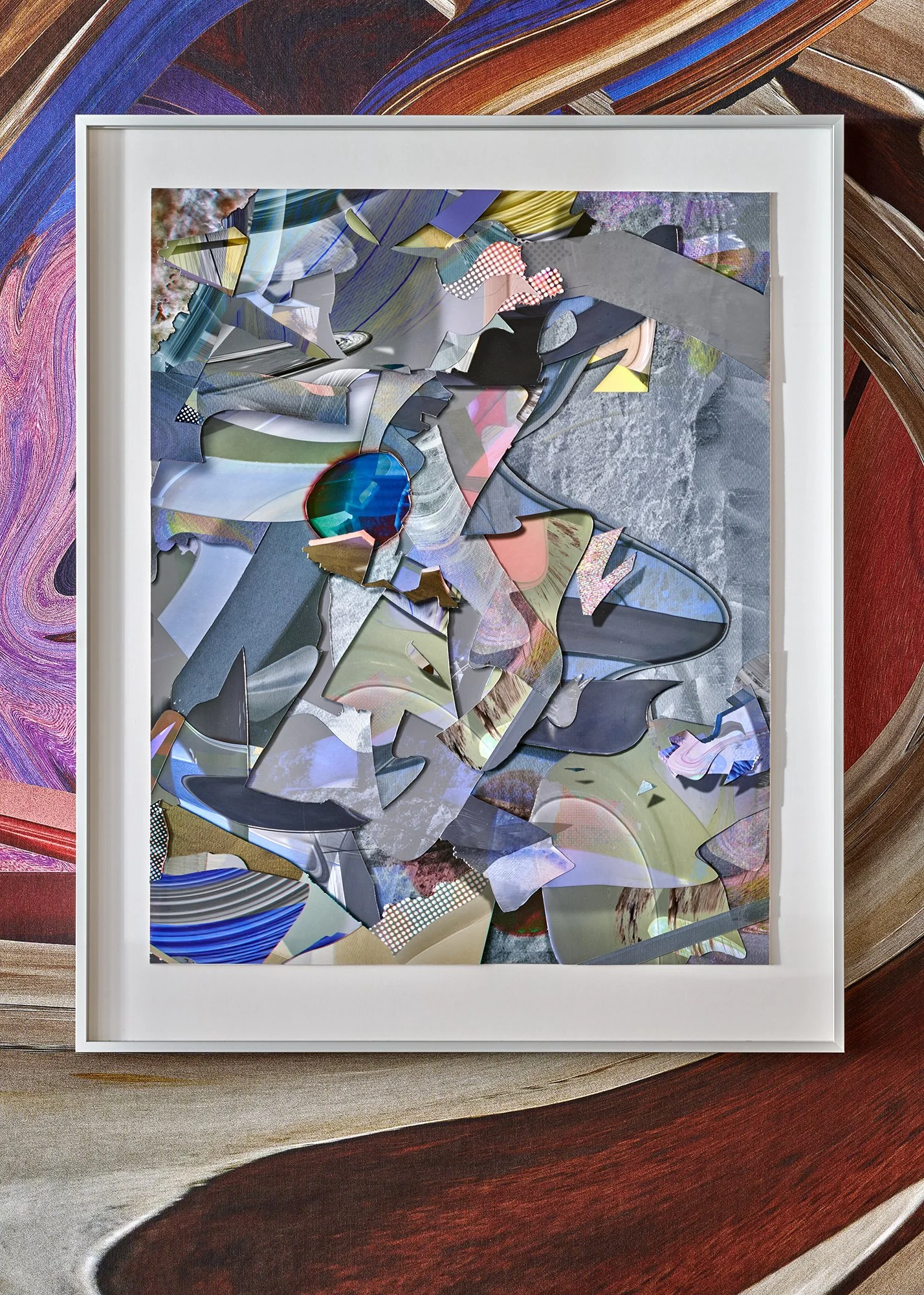 An abstract collage artwork featuring a variety of colorful paper and material pieces arranged inside a white frame, placed on a wooden surface with reddish and brown tones.