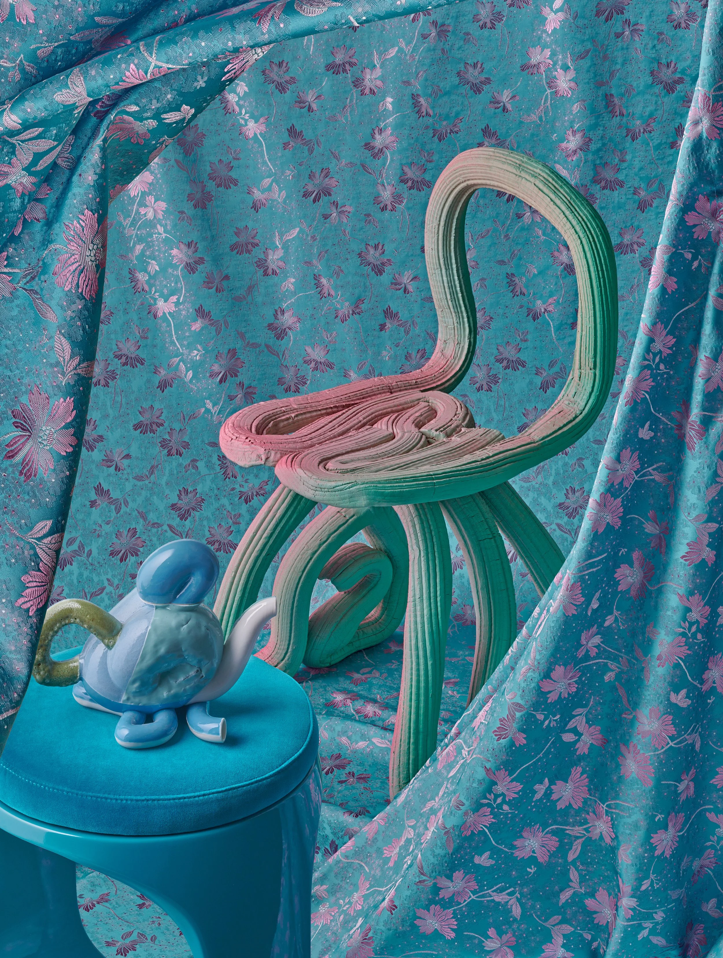 A surreal scene with a cloth backdrop covered in pink and purple floral patterns, featuring a wooden chair with a whimsical, curvy design, and a decorative blue elephant figurine on a matching blue stool.