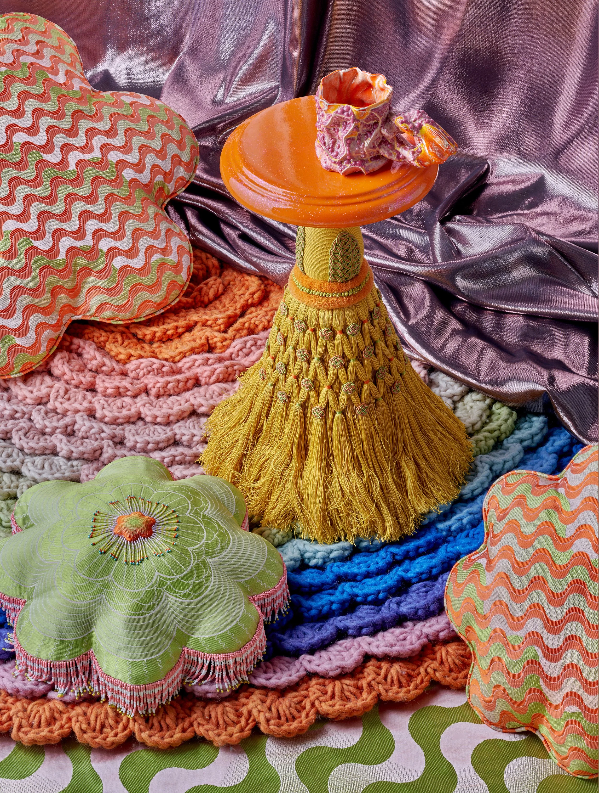 Colorful decorative objects including a yellow fringed skirt with beaded details, a gray ceramic vase on an orange dish, plush fabric and crochet blankets, and patterned cushions with abstract wave and floral designs, arranged on a multicolored textu