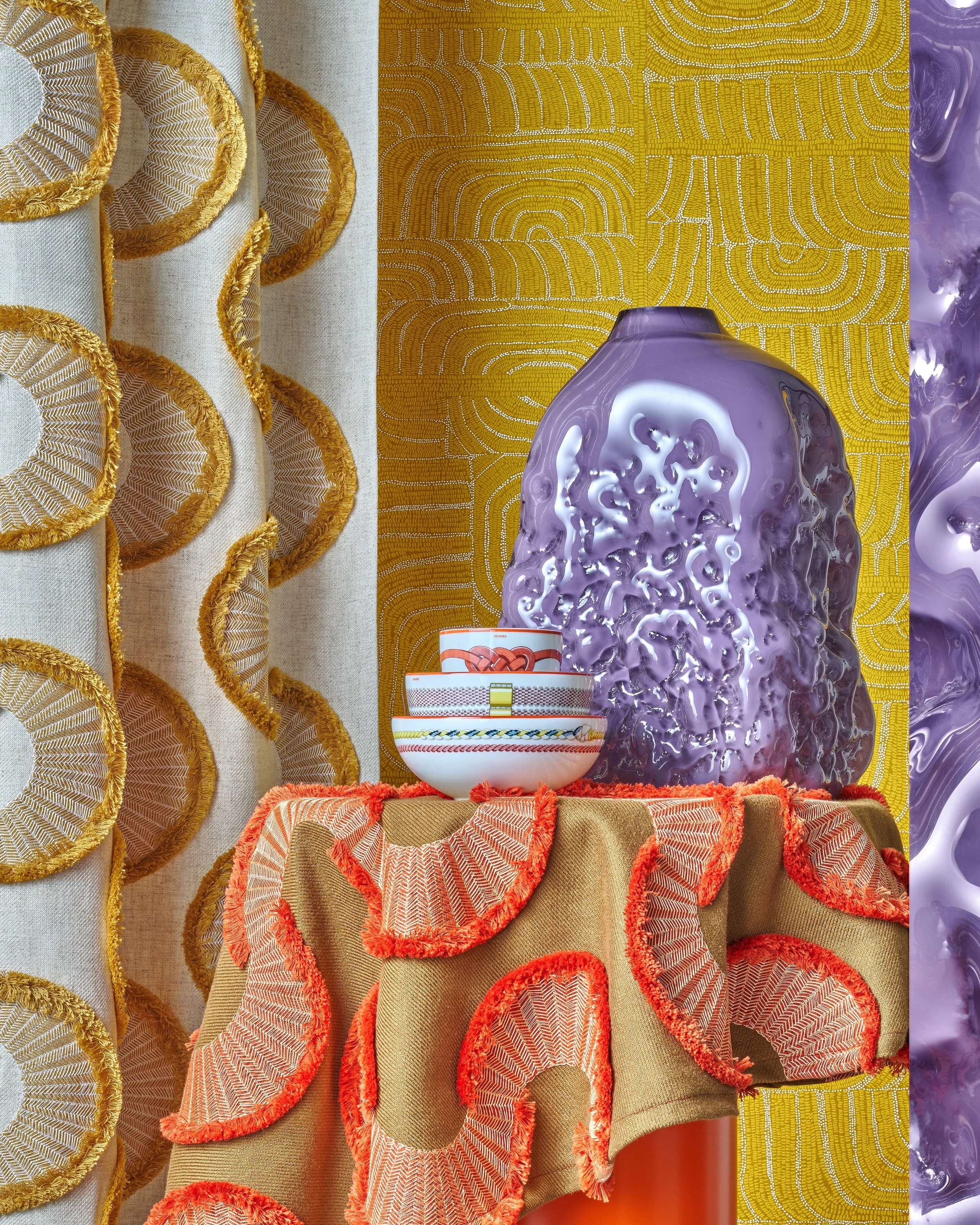 Vase with textured purple surface, stacked bowls with colorful patterns, and fabric with circular embroidery and fringes, all on a table with a patterned yellow backdrop.