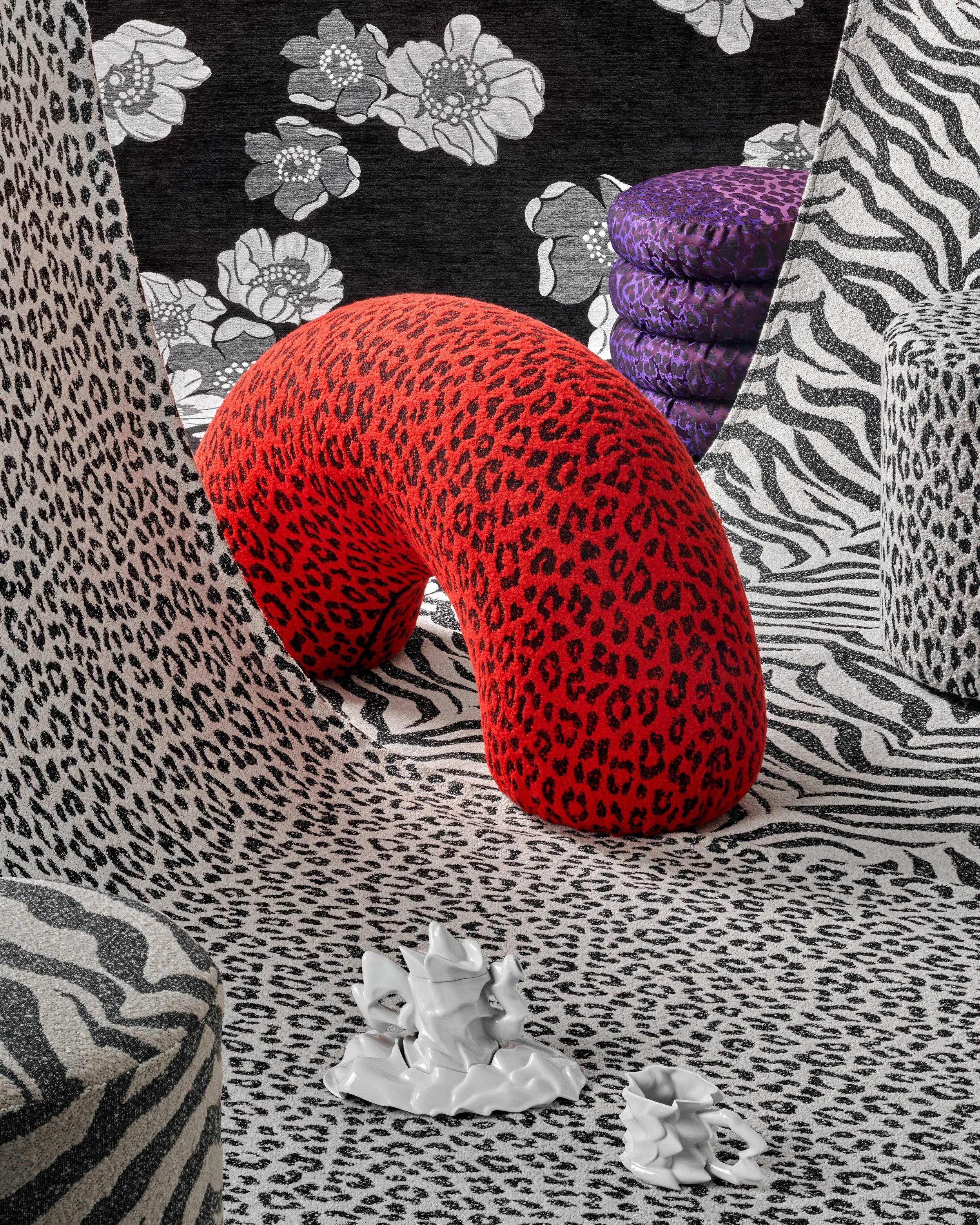 An interior with animal print patterns on furniture and walls, including a large curved cushion with a red and black leopard print, a purple animal print pillow, and a black and white zebra print wall. There are two white crumpled ceramic vases or sc