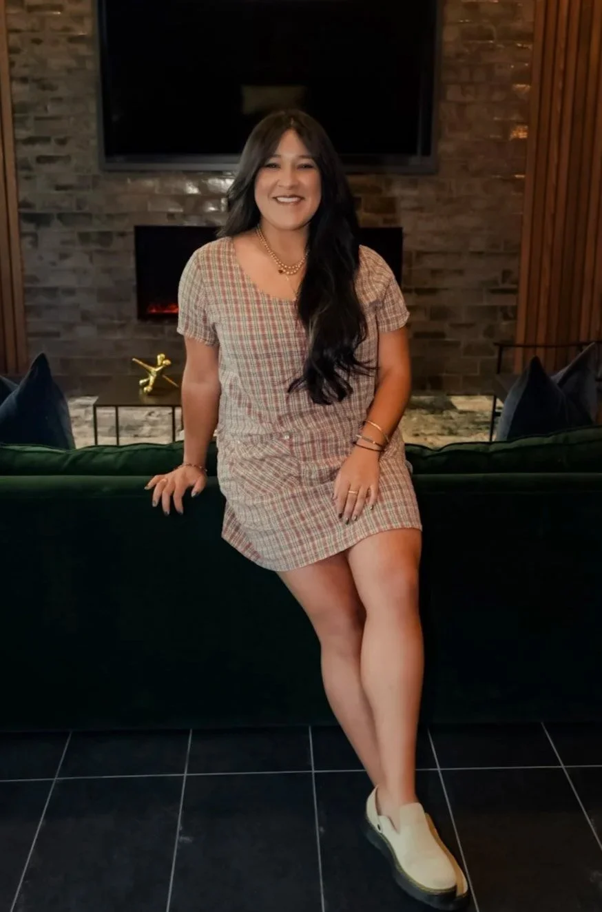 A woman with long dark hair sitting on a dark green sofa in a cozy room with brick walls and a fireplace, smiling at the camera.