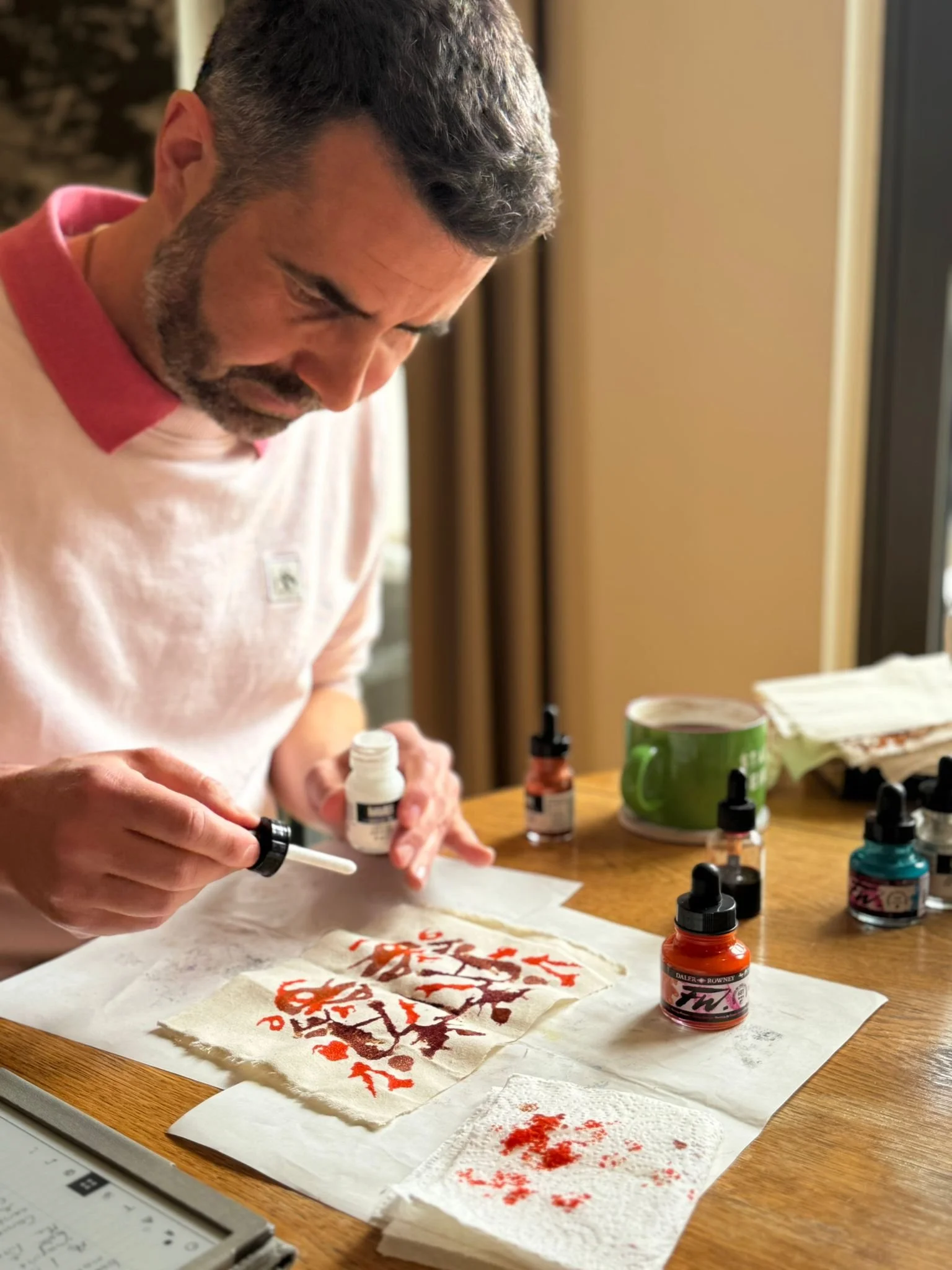 Alex is shown in the middle of making a Talisman.  He holds a pot of white ink and a dropper over the sheet of calico.  The calico shows the earlier words and colours that form the Talisman.  Other bottles of ink are scattered around the table.