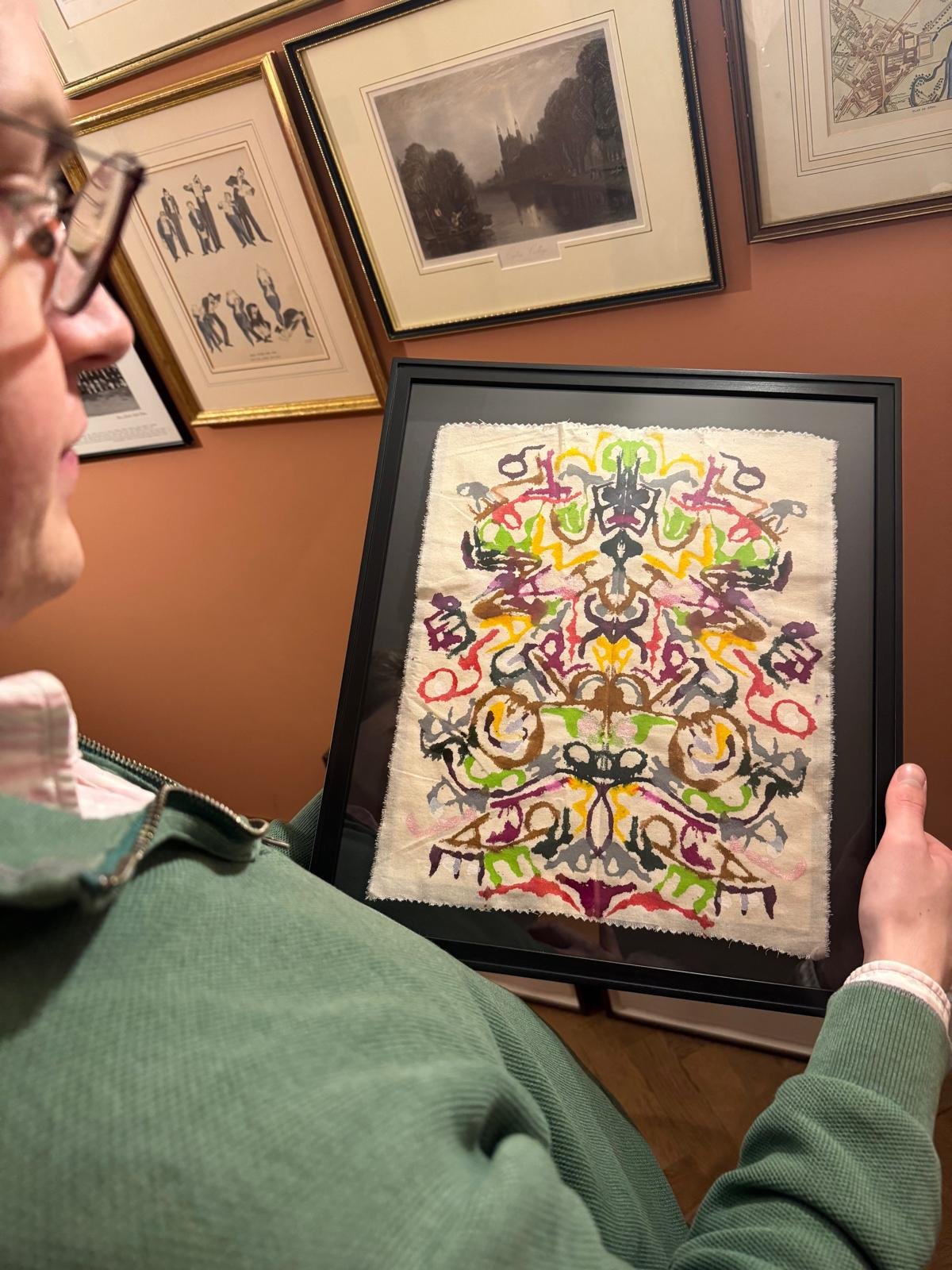 A man holds a framed Talisman in his hands, with other framed photos & art in the background.  The Talisman is made of many colours, all blotted to create the final piece.  This is a couple Talisman using words of meaning from their relationship.