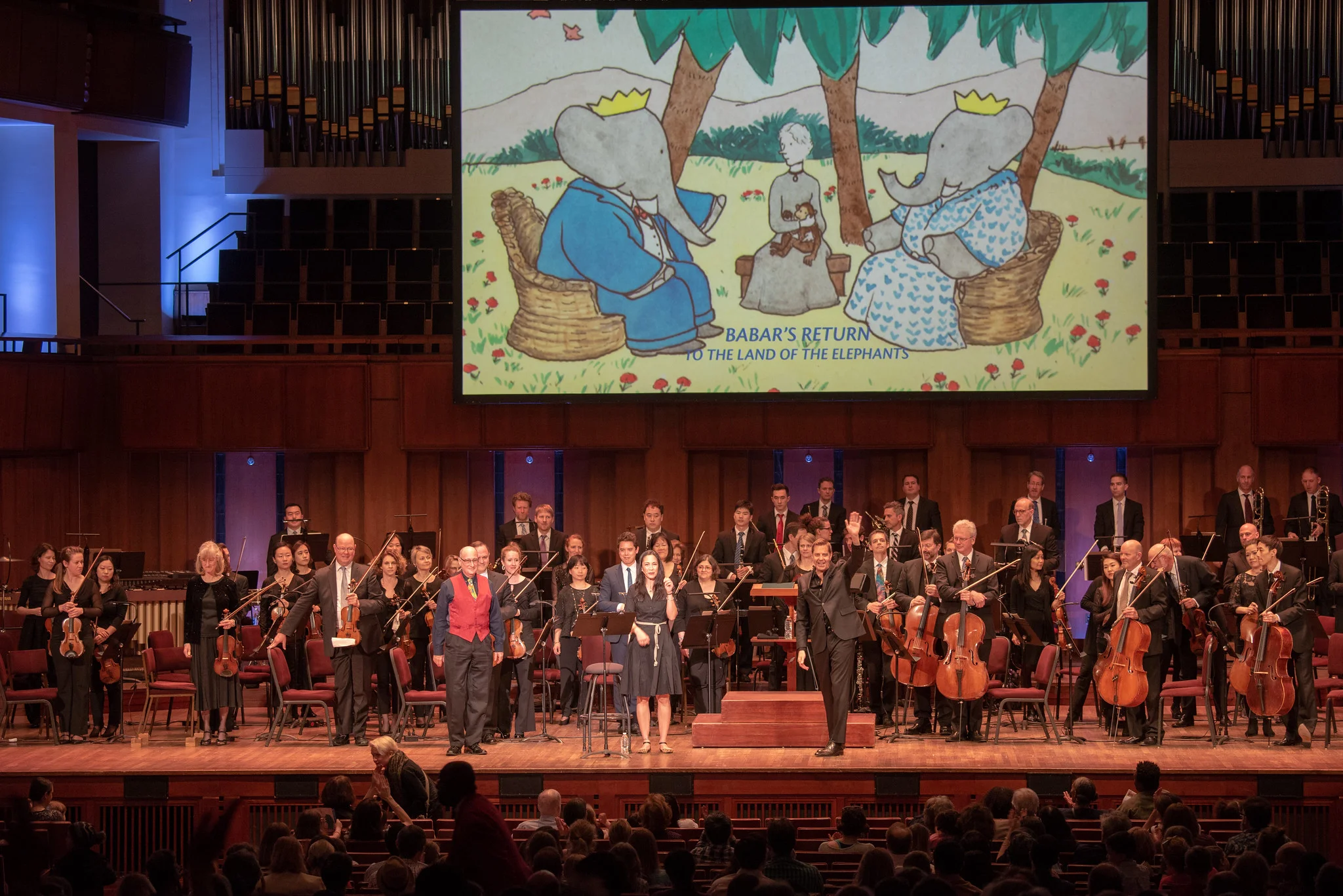(l-r in front): Composer Raphael Mostel, narrator Regina Aquino, conductor Steven Reineke, bows after first U.S. performance, Babar’s Return to the Land of the Elephants National Symphony Orchestra, John F Kennedy Center for the Arts, Washington, D.C., April 7, 2019 Photo: Erica Bruce (full size photo available). Multimedia image: original Jean de Brunhoff watercolor, Kendra & Allan Daniel Collection. Used with permissions