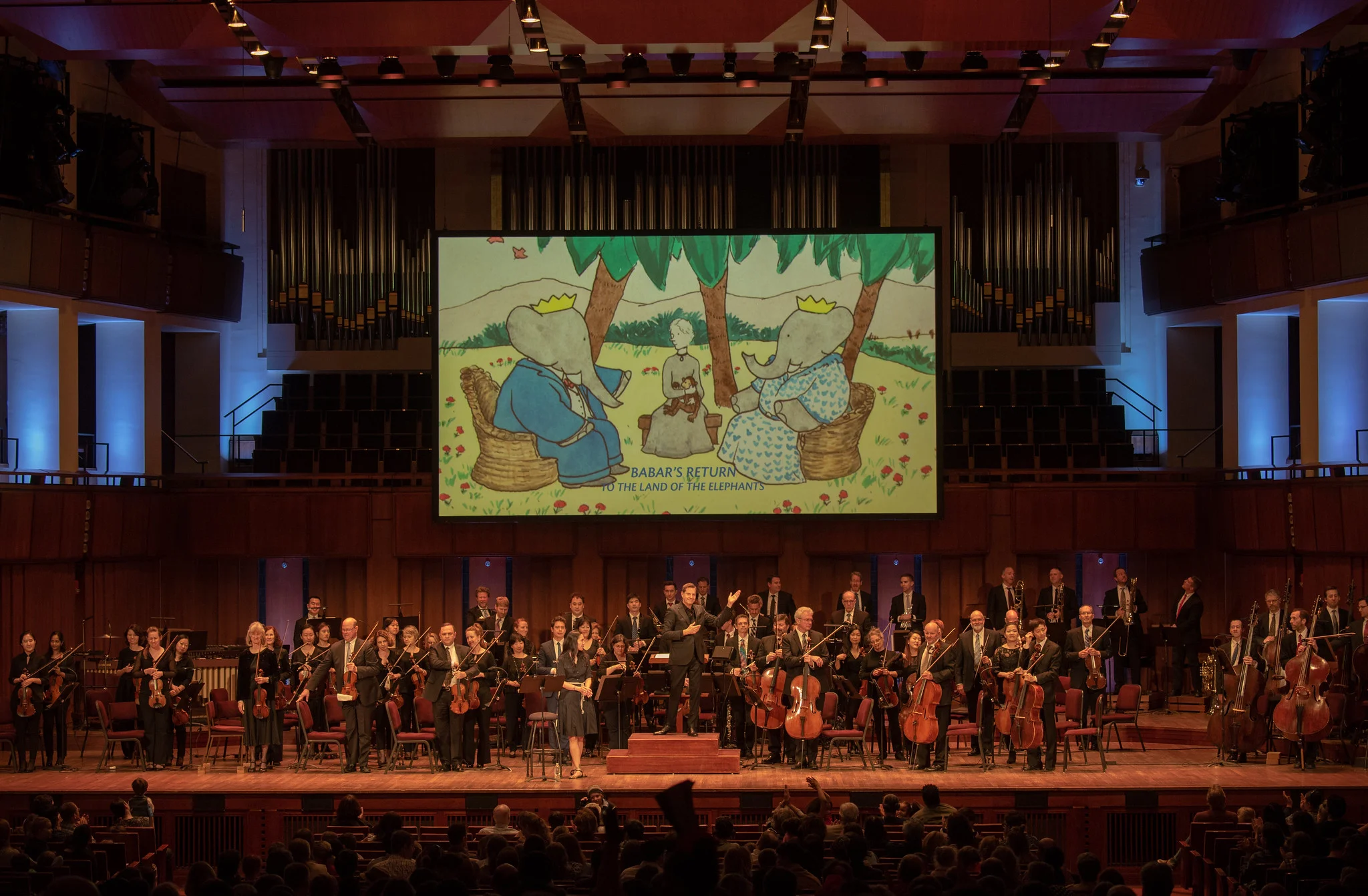 First U.S. performance, Raphael Mostel / Jean de Brunhoff Babar’s Return to the Land of the Elephants for orchestra. Narrator Regina Aquino, conductor Steven Reineke with National Symphony Orchestra, John F Kennedy Center for the Arts, Washington, D.C., April 7, 2019 Photo: Erica Bruce, for Mostel.com (full size, hi-res photo available). Multimedia image: Jean de Brunhoff watercolor from the Kendra and Allan Daniel Collection, Used with permissions
