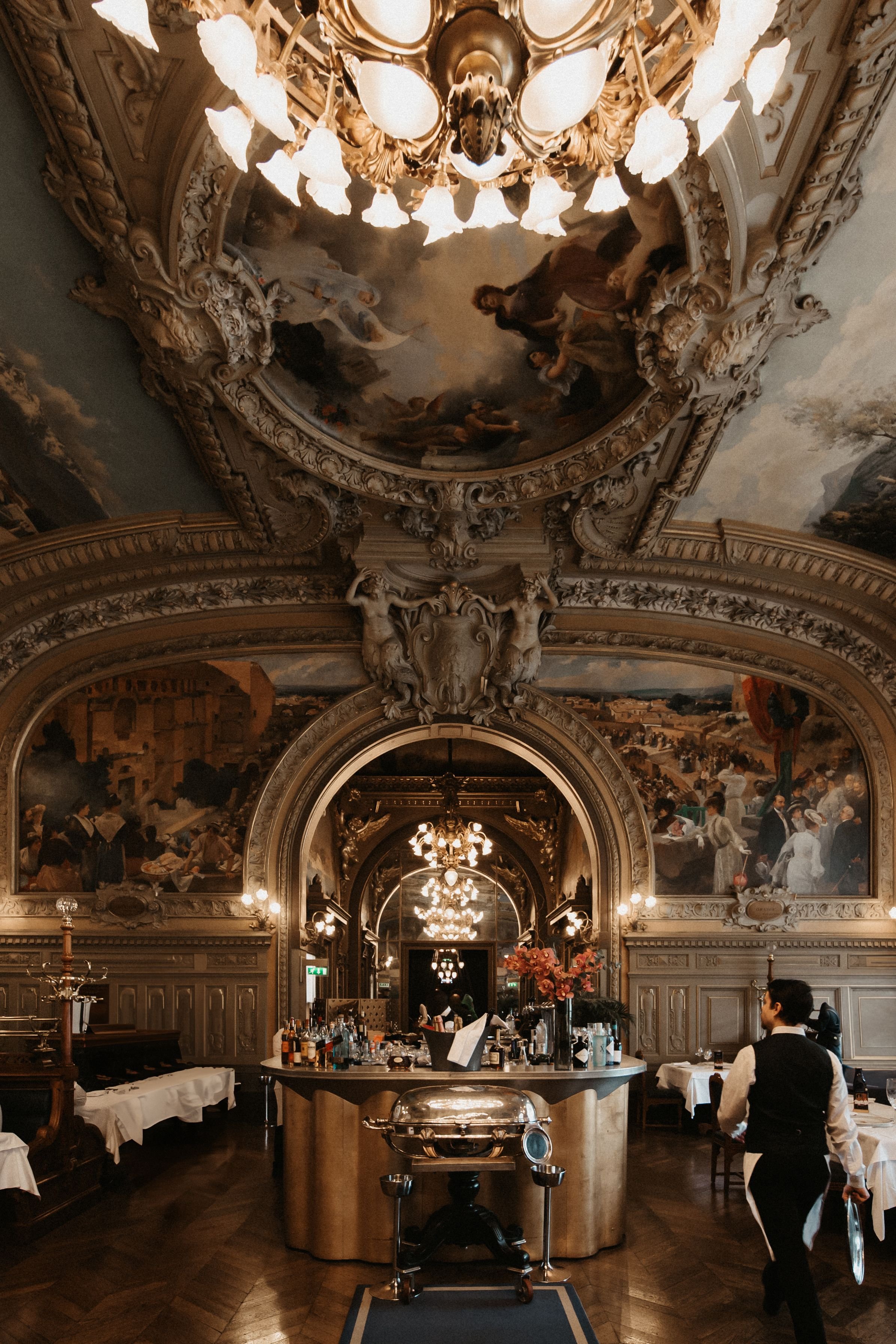 Elegant restaurant interior with a high ornate ceiling featuring a large chandelier and classical ceiling painting, a bar with bottles and glasses, and waitstaff preparing tables.