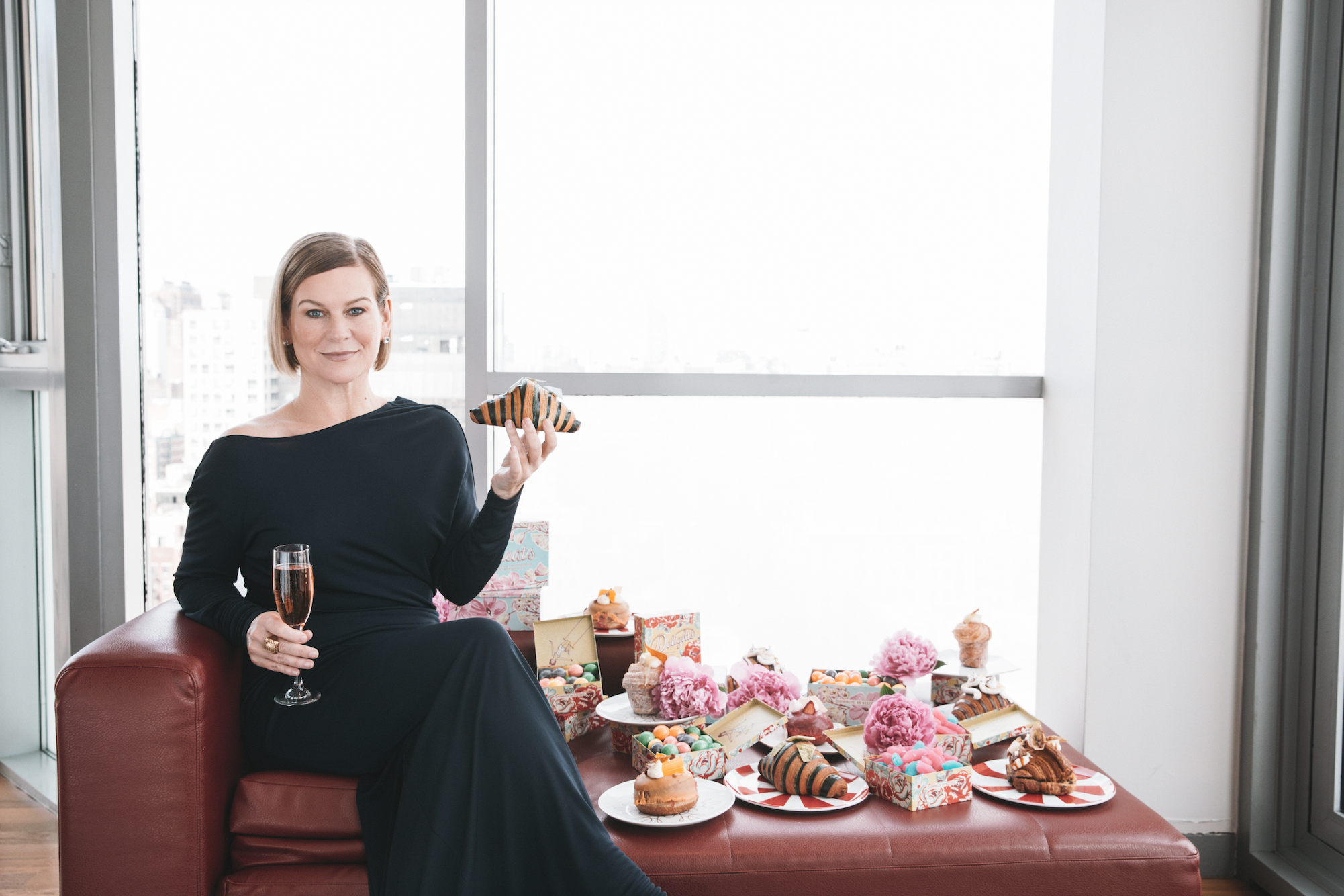 A woman in a black dress sitting on a red couch holding a glass of wine and a slice of cake, surrounded by colorful desserts and pink flowers inside a modern high-rise apartment with large windows.