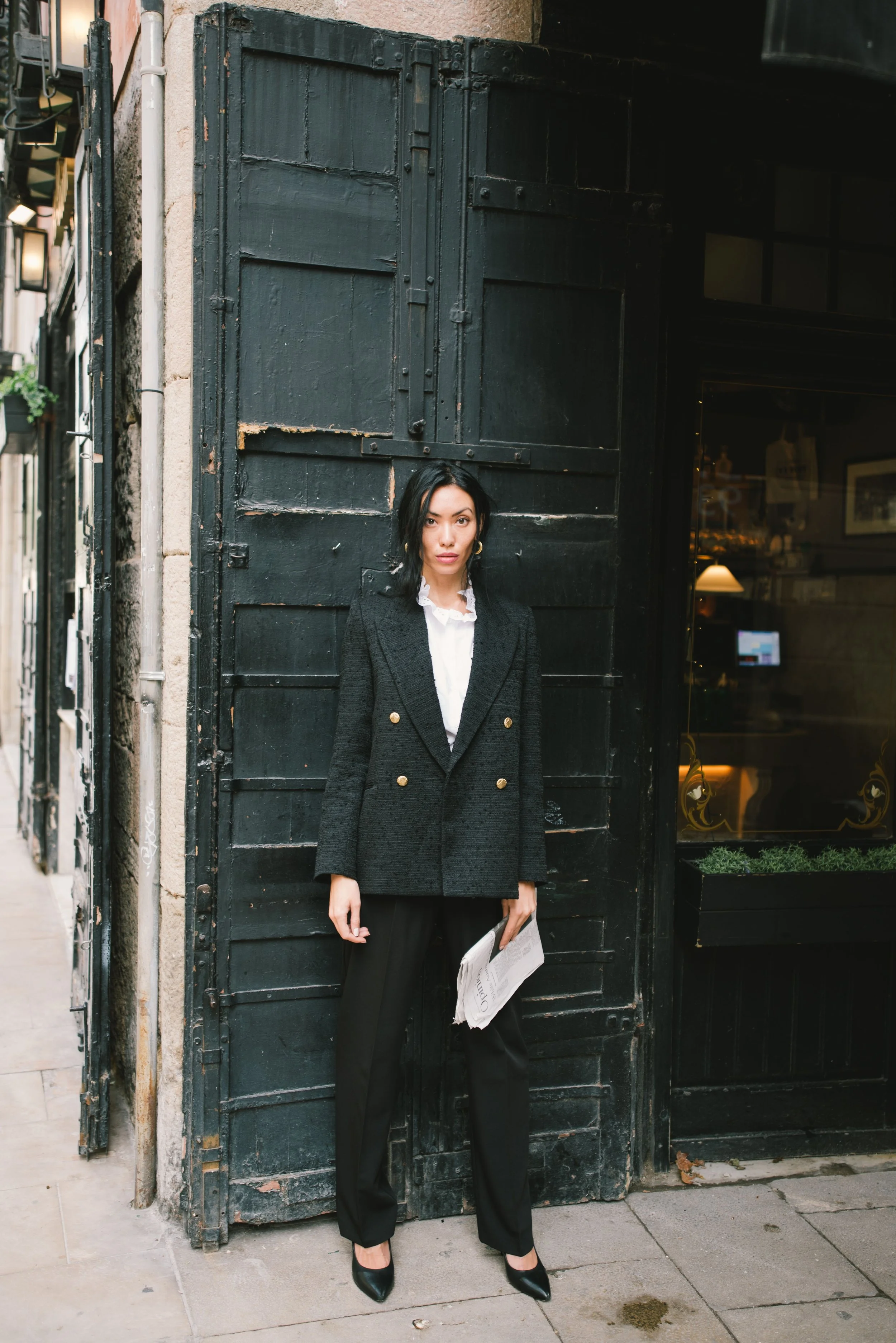 A woman standing on a city sidewalk in front of a large black wooden door. She is wearing a black blazer with gold buttons, black pants, and black heels, holding a newspaper.