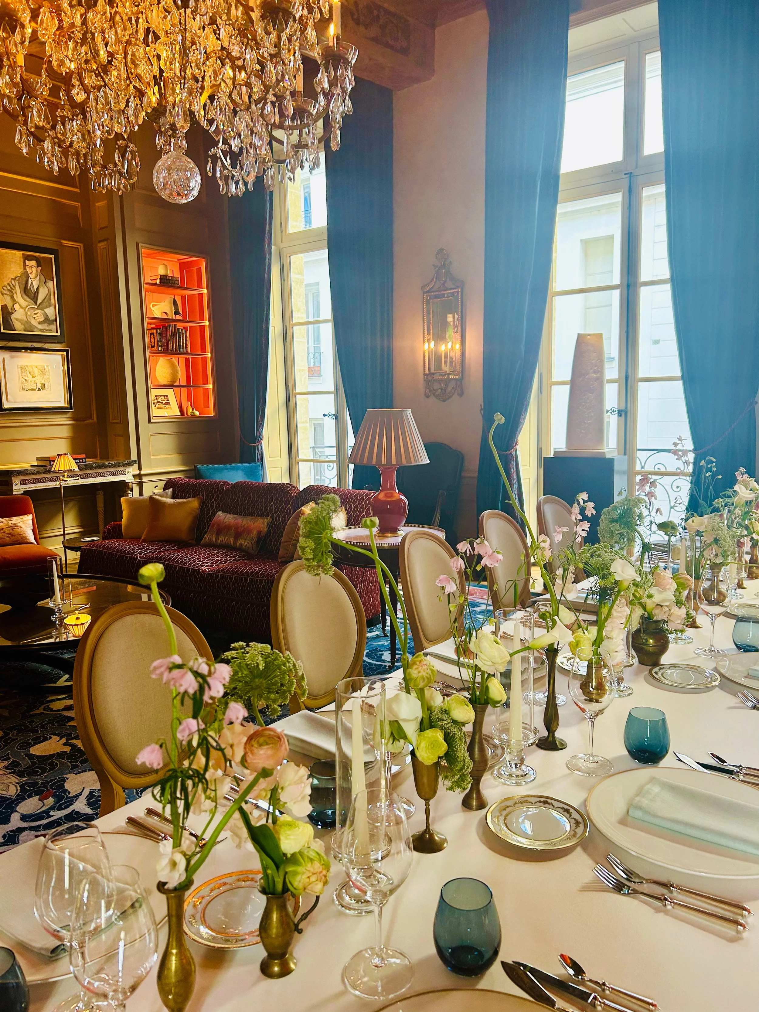 Elegant dining room with a large chandelier, tall windows with blue curtains, and a lavish table set with floral arrangements, fine china, and glassware.
