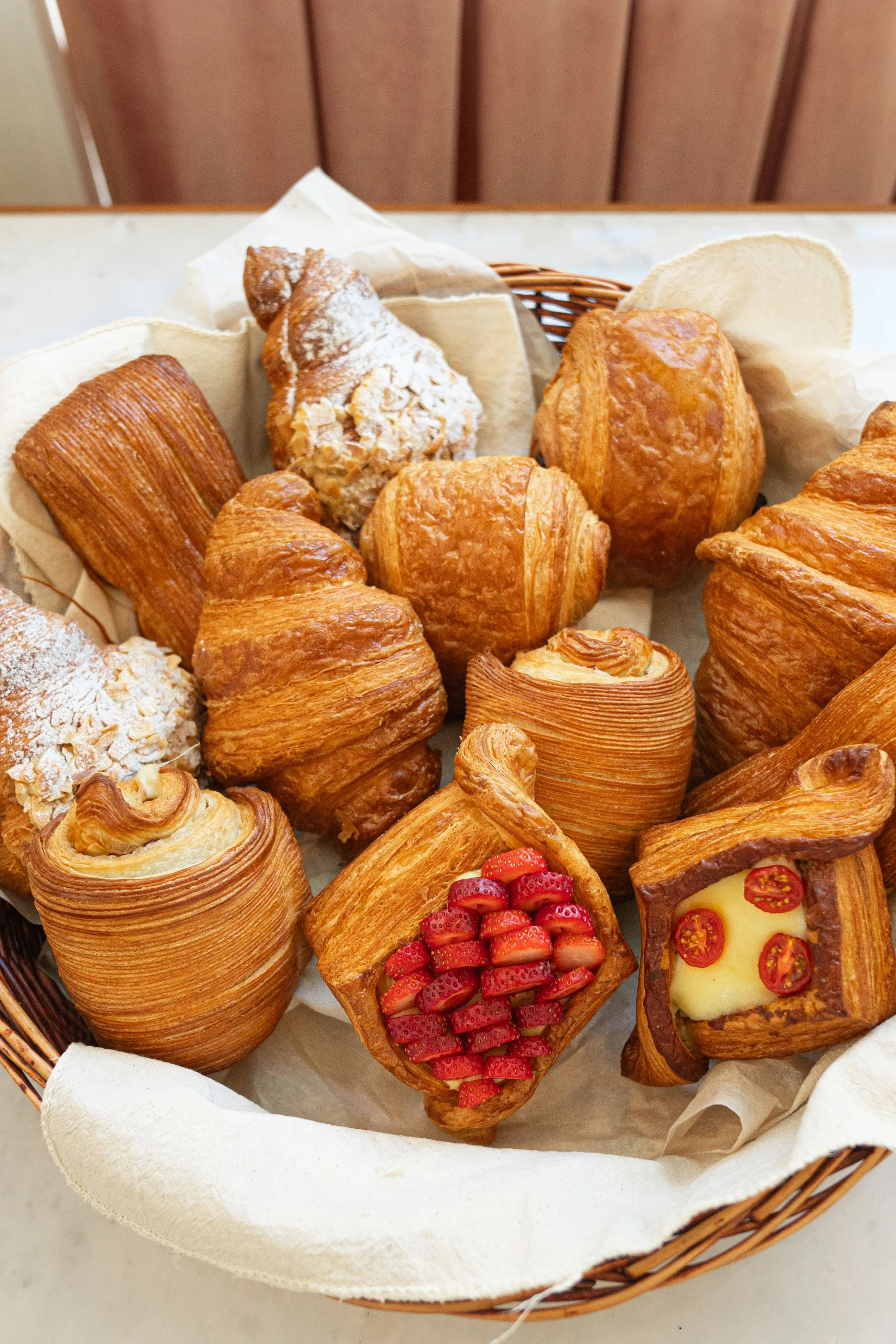A basket filled with assorted pastries including croissants, Danish pastries topped with strawberries, and other flaky baked goods, all arranged on white parchment paper.