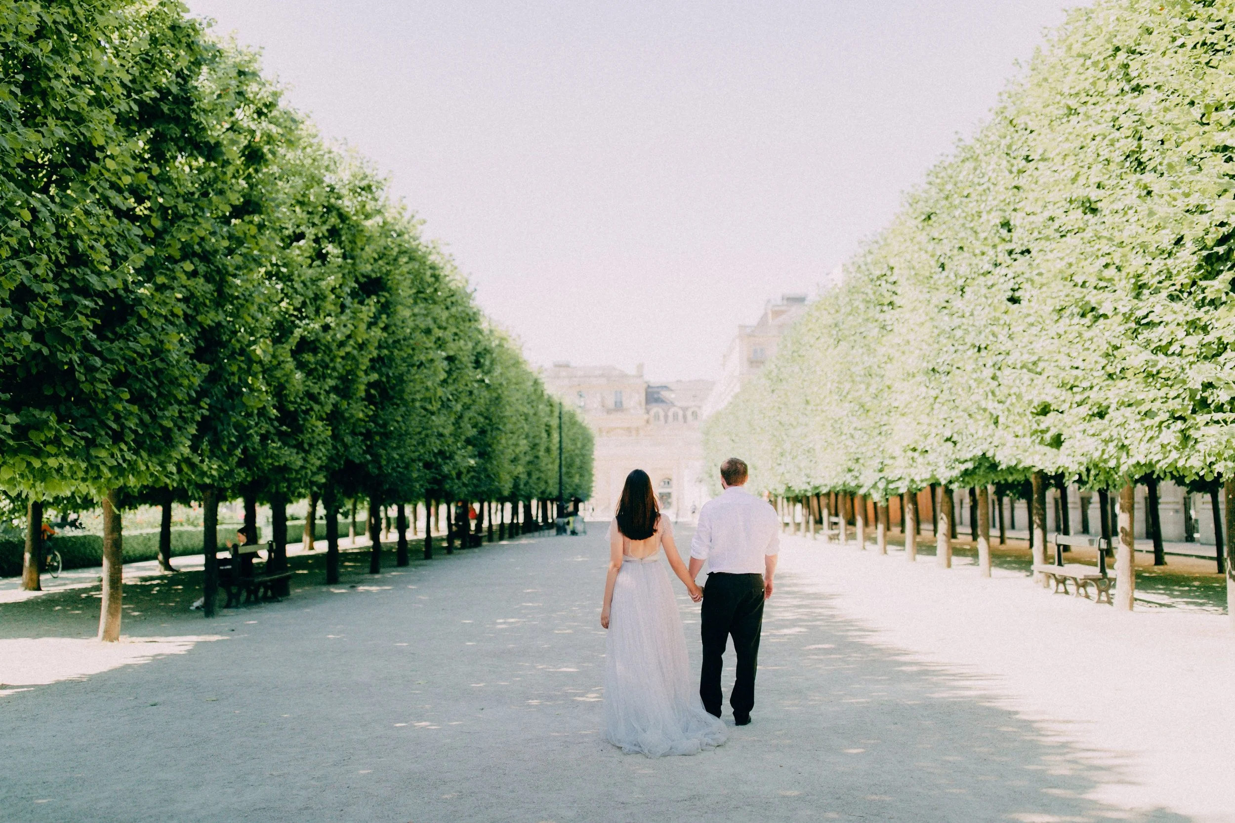 A couple walking hand in hand in a park, surrounded by lush green trees, with a bright sky overhead.