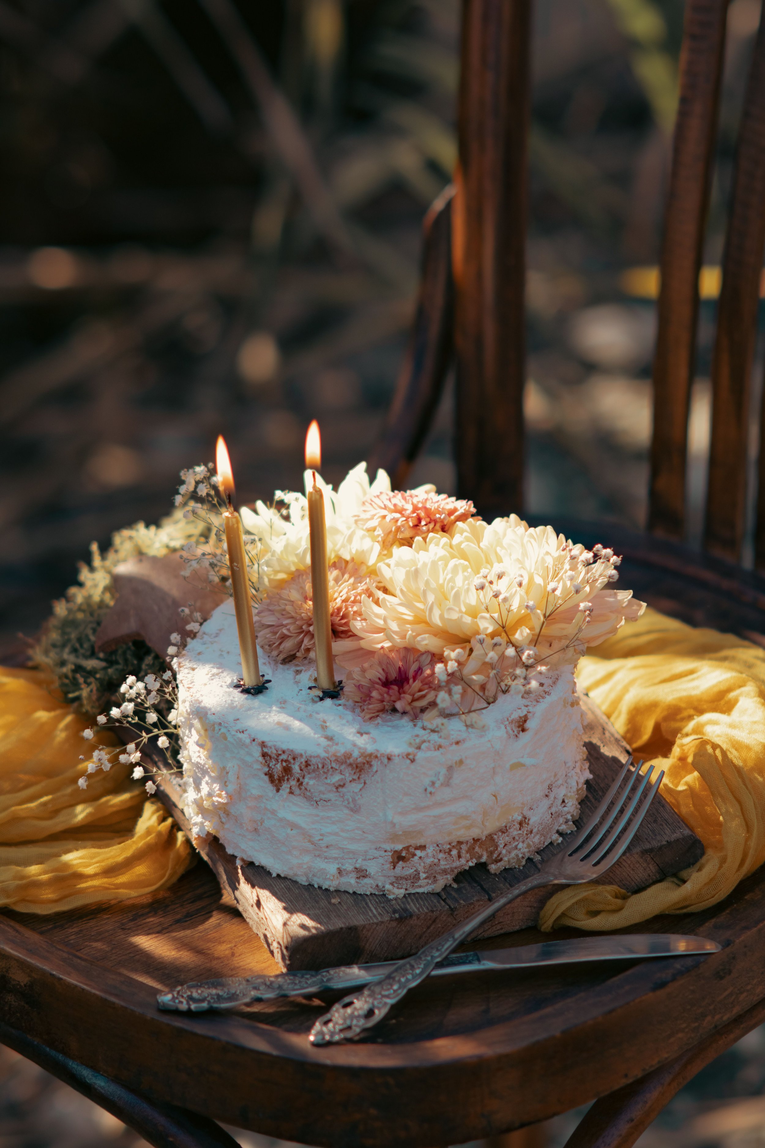 A white birthday cake decorated with pink and white flowers, two lit candles, and small wisps of baby's breath flowers, placed on a wooden surface with a yellow cloth underneath, outdoors with natural outdoor lighting.